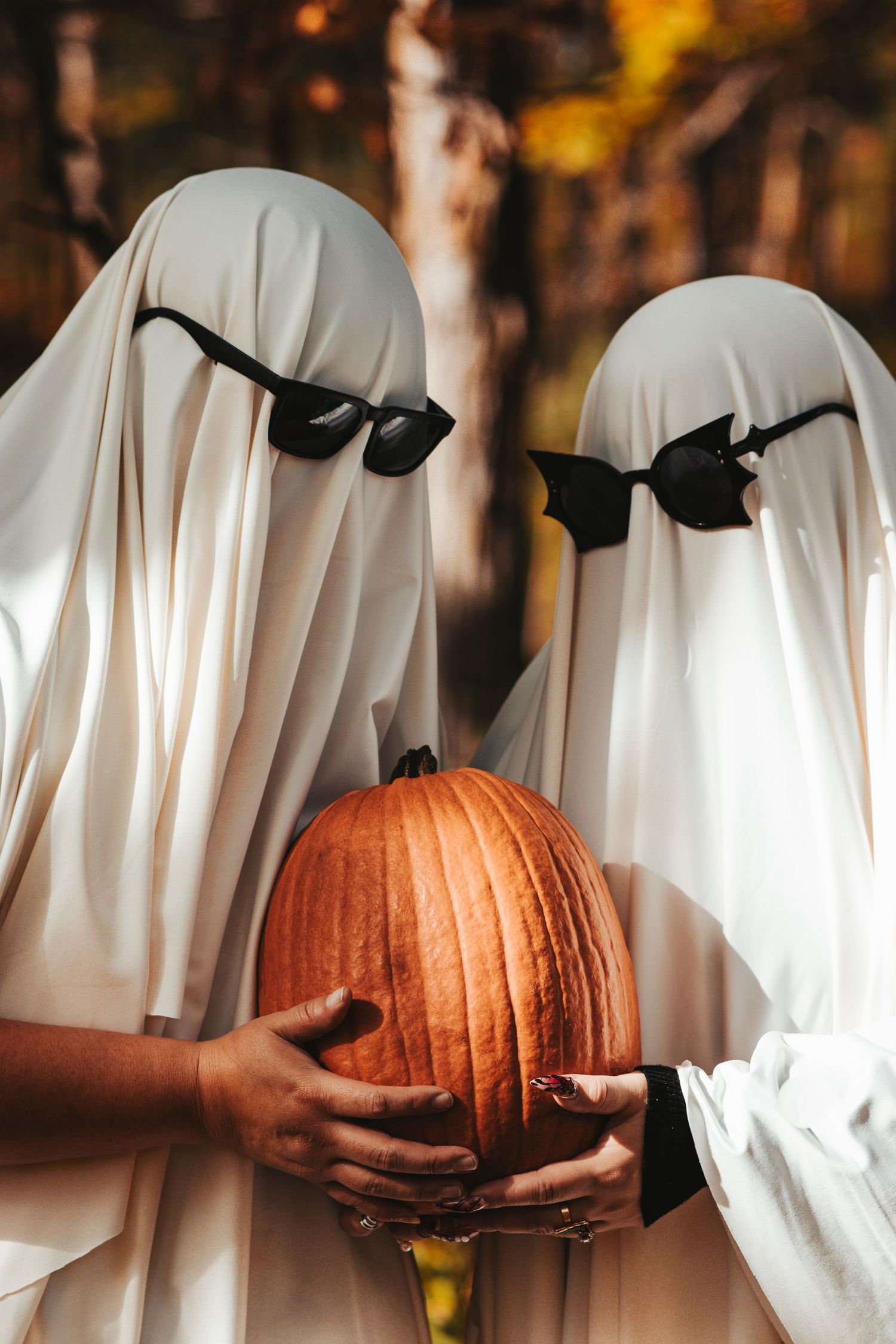 Two ghost costume figures in sunglasses hold a pumpkin in an autumn forest setting.