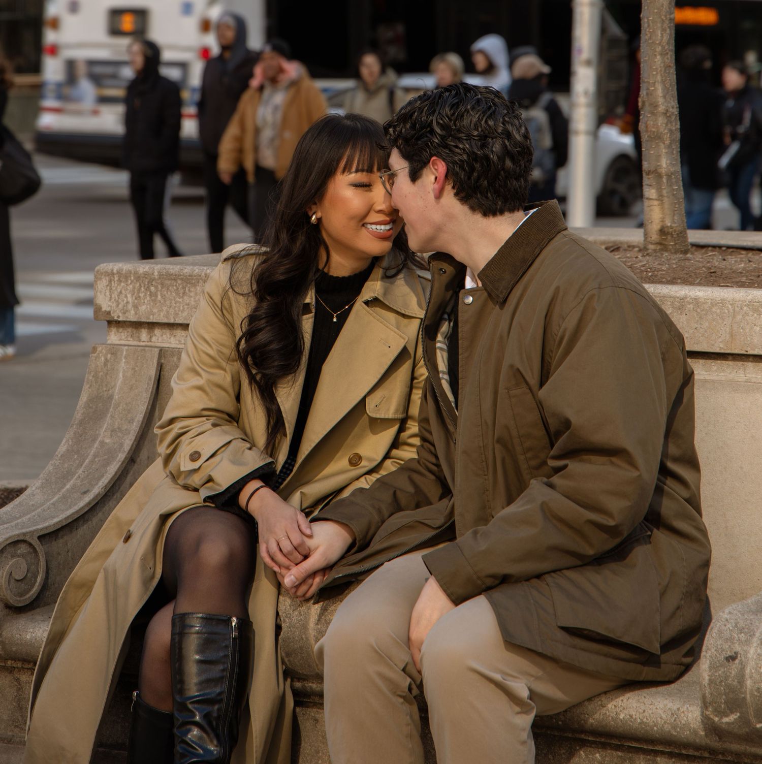 Happy couple sits close together on a stone bench on a sunny city street.
