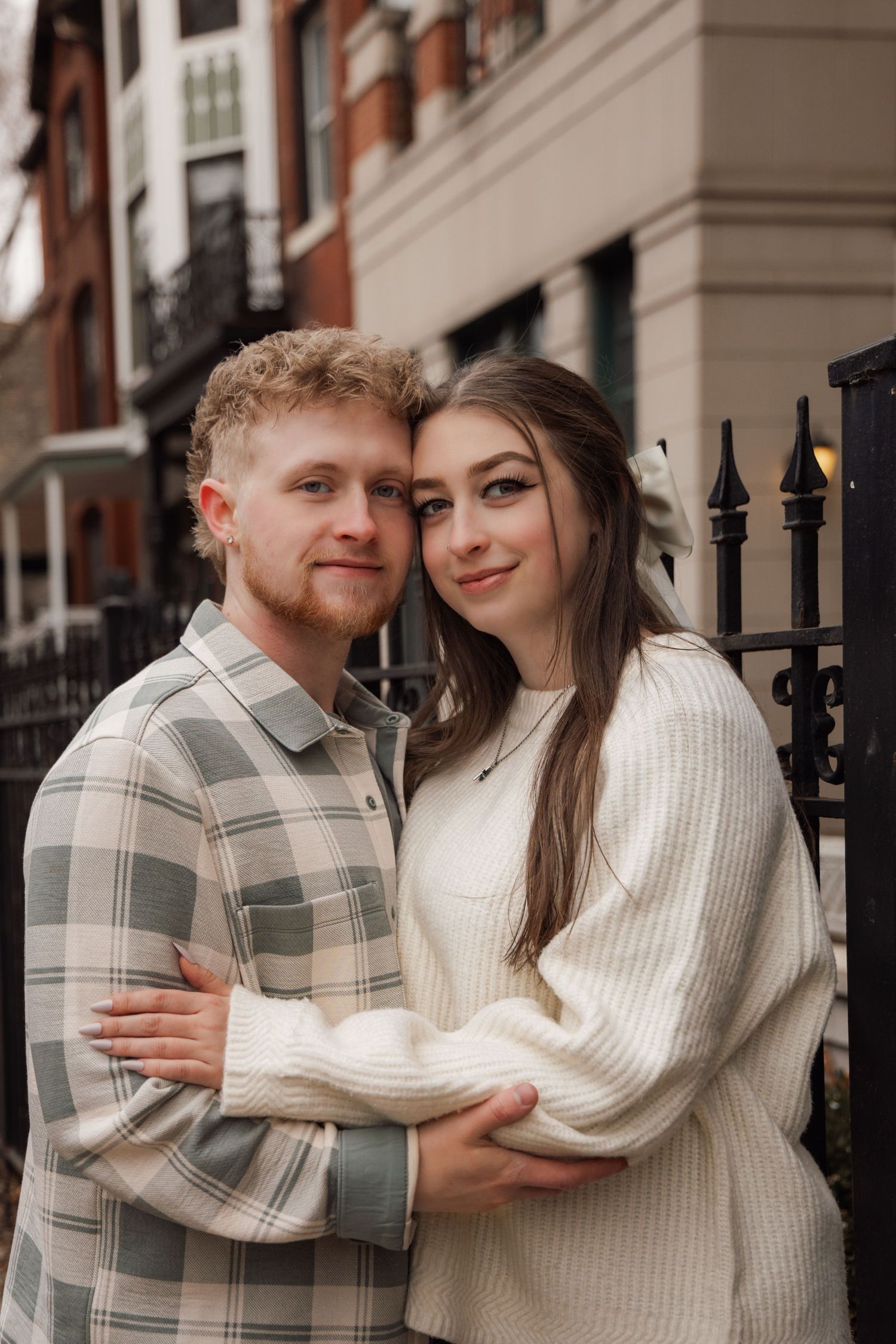 Young couple embracing and smiling by black iron fence with brick townhouses in background.