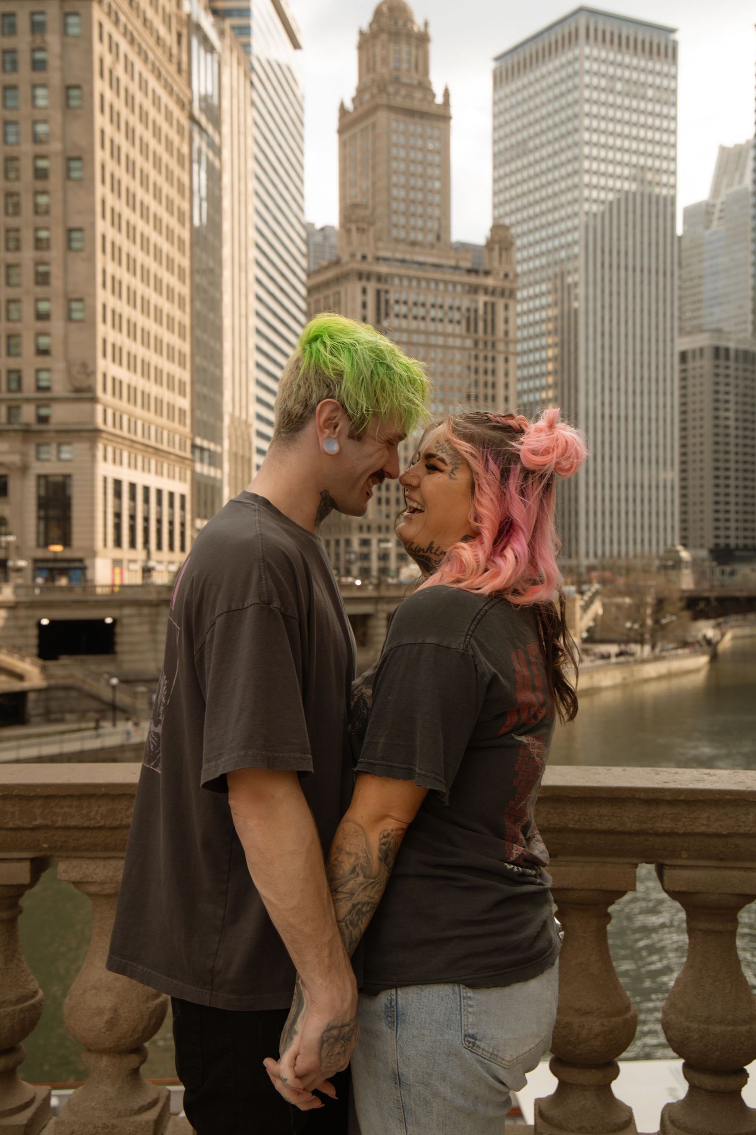 A couple with colorful hair holds hands and smiles at each other on a bridge with Chicago skyline behind them.
