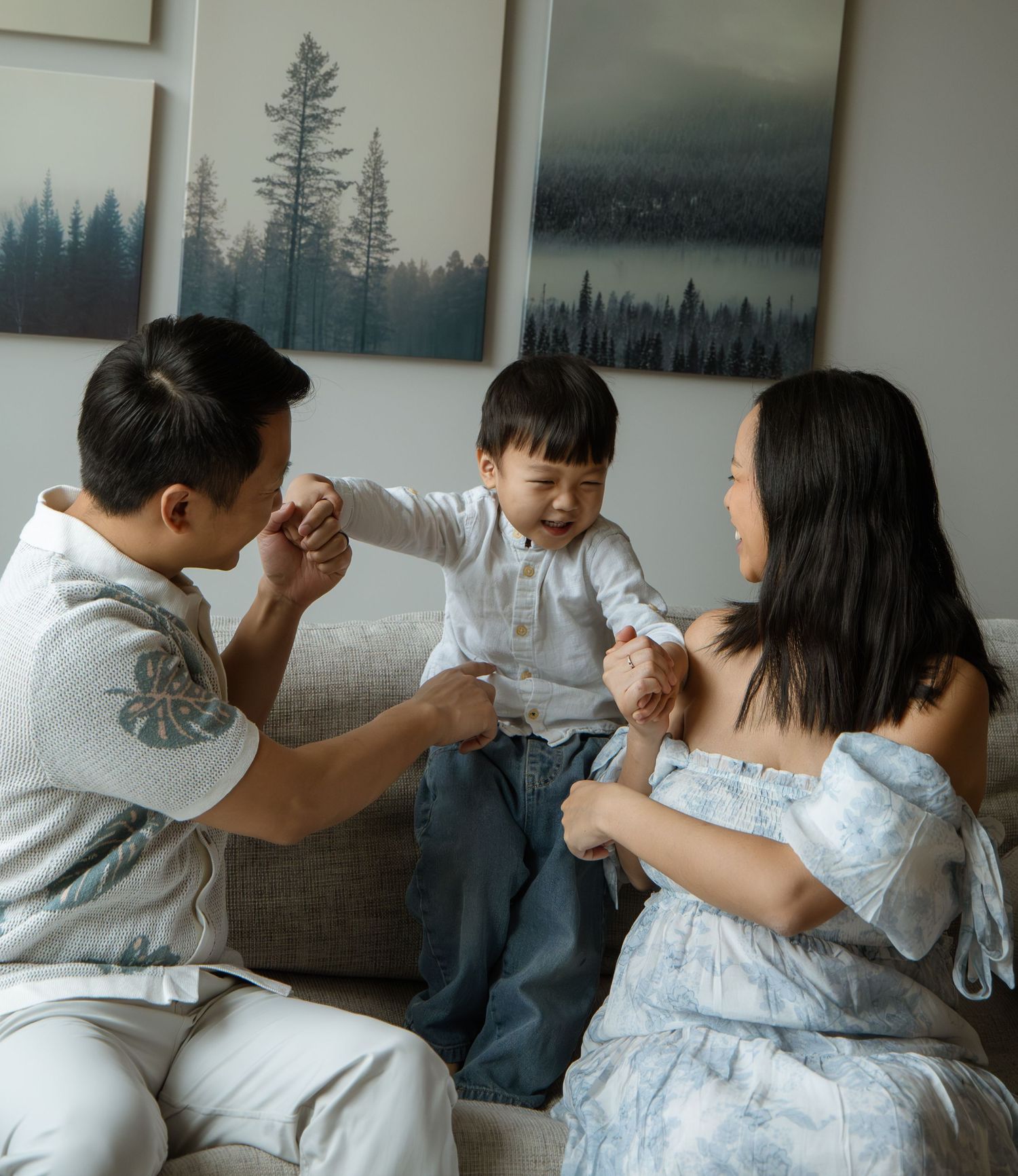 A happy Asian family with a young toddler boy sitting on a couch, laughing and holding hands together at home.