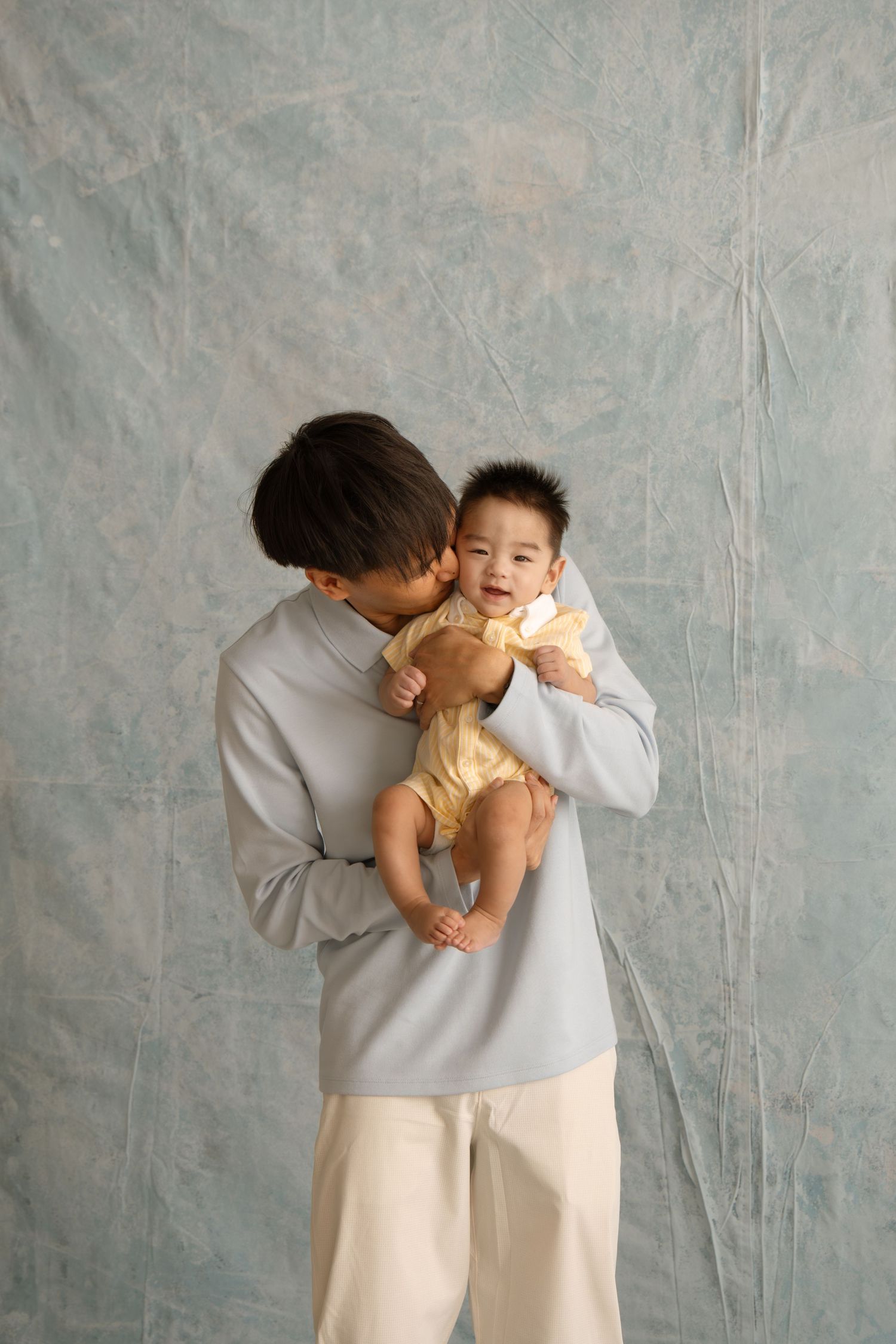 A parent in a light blue sweater tenderly kisses a smiling baby in a yellow outfit against a gray backdrop.