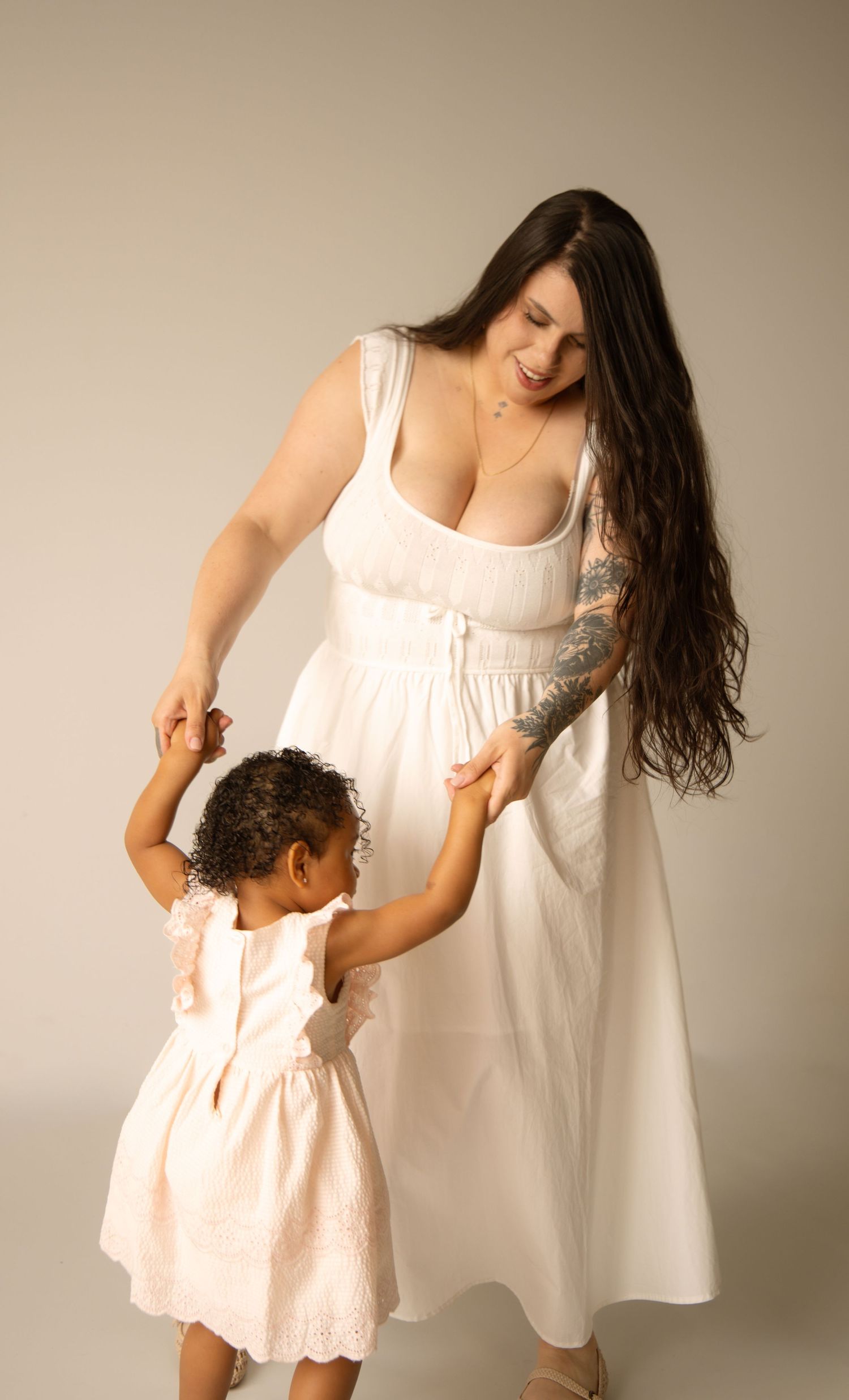 A woman in a white dress dances with a young girl in a pink dress in a studio photo shoot.