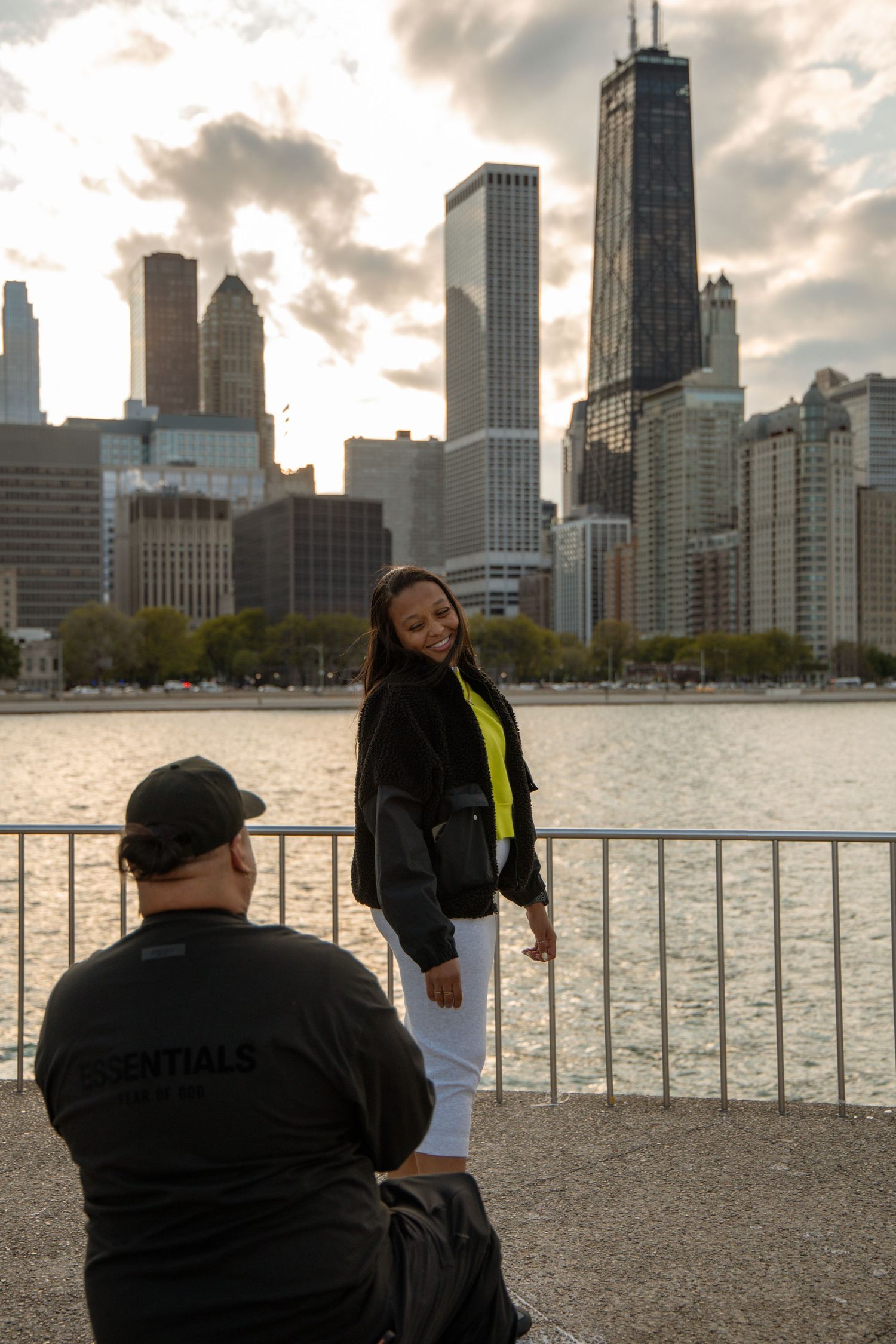 Woman poses by Chicago waterfront railing with city skyline at sunset while photographer captures her portrait.