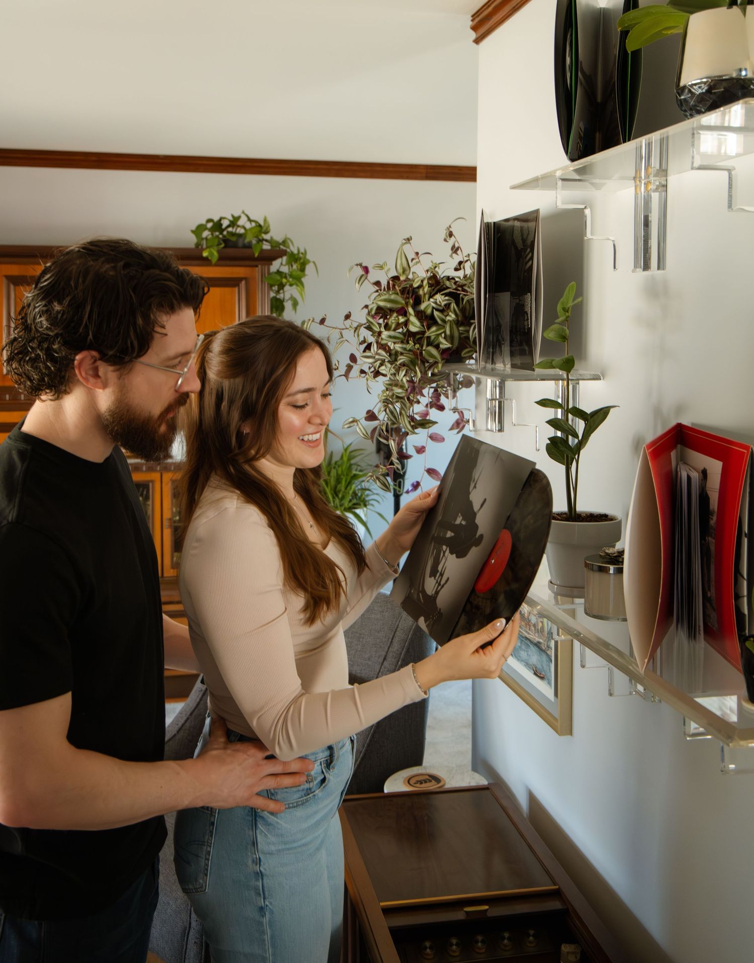 Couple smiling and browsing vinyl records on a wall-mounted shelf in a cozy home living room.