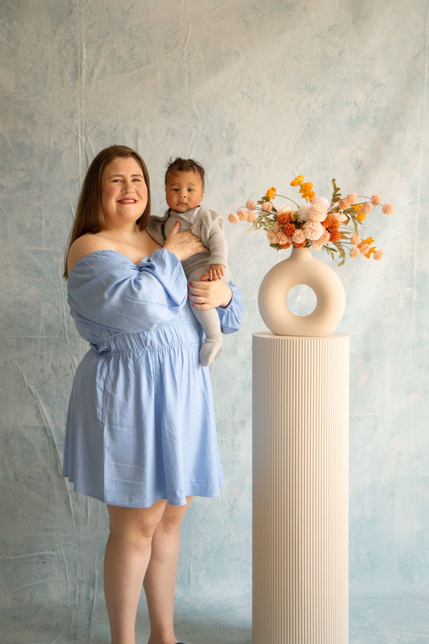 A smiling woman in a blue off-shoulder dress holds her baby near a white vase with orange flowers on a pedestal.