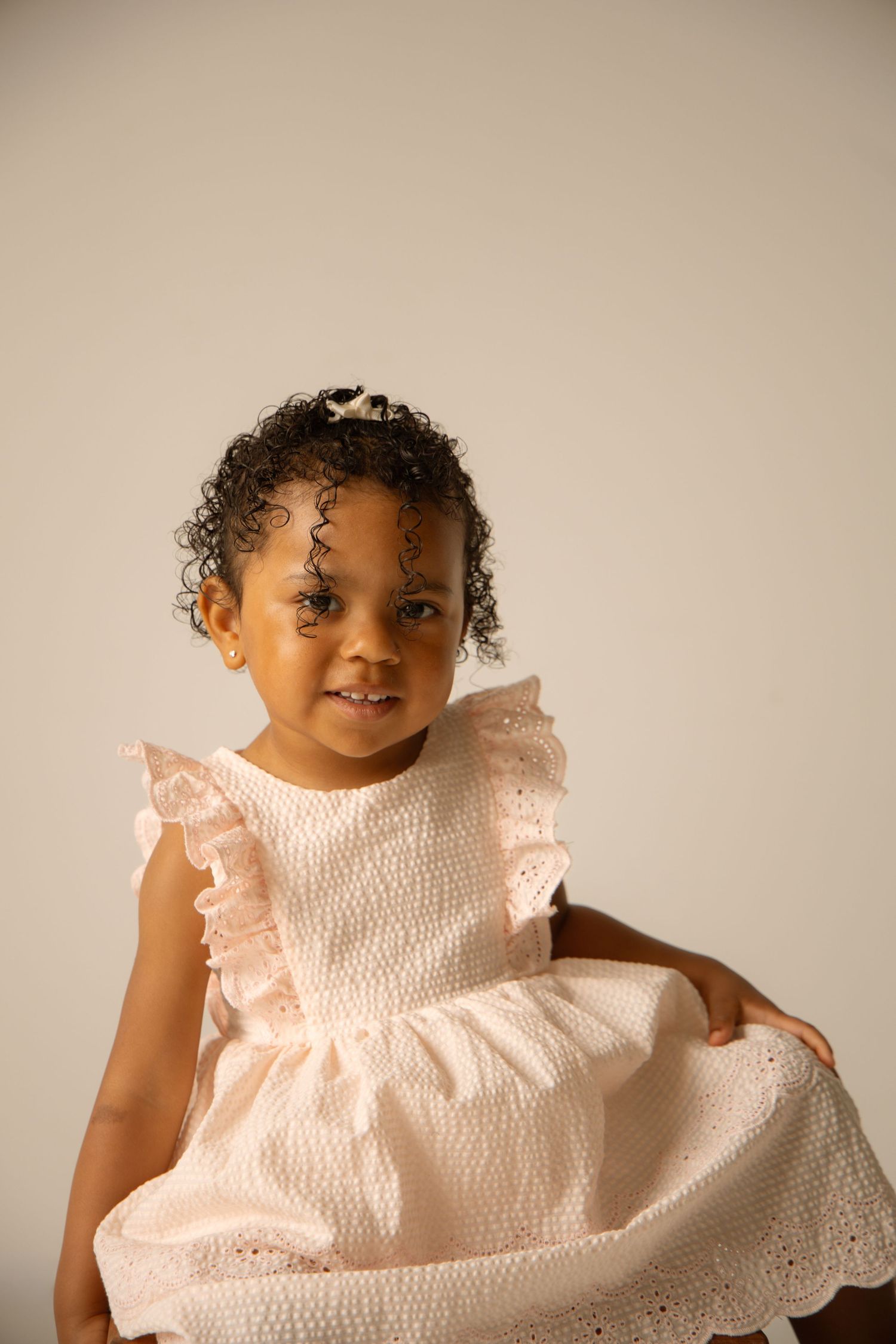 A young girl with curly hair smiles in a light pink ruffled dress against a neutral background.