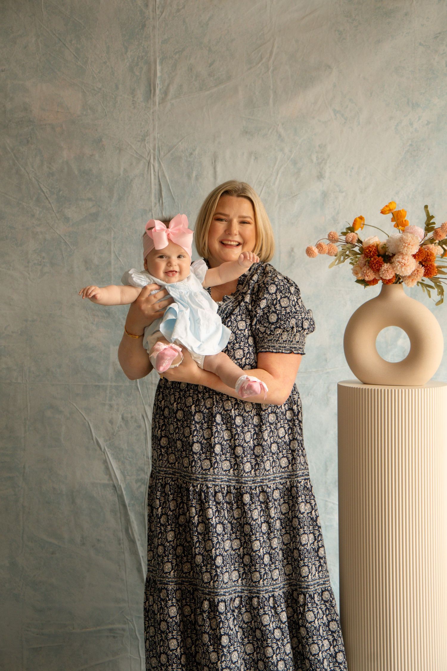 A smiling woman in a floral dress holds a baby girl with a pink bow near a vase of orange flowers.