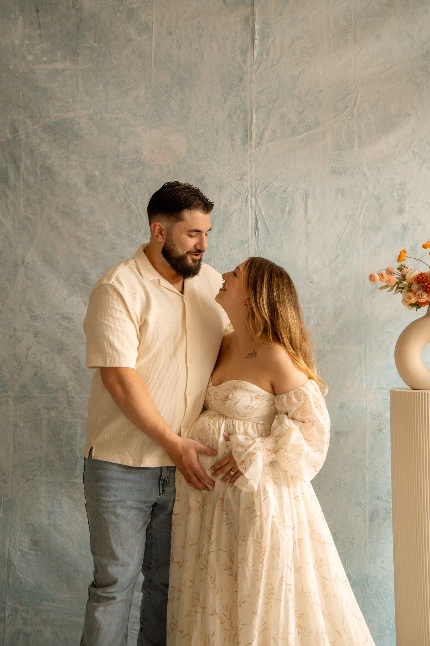 A couple shares a tender moment, the pregnant woman in a lace gown held by her bearded partner in a soft studio setting.