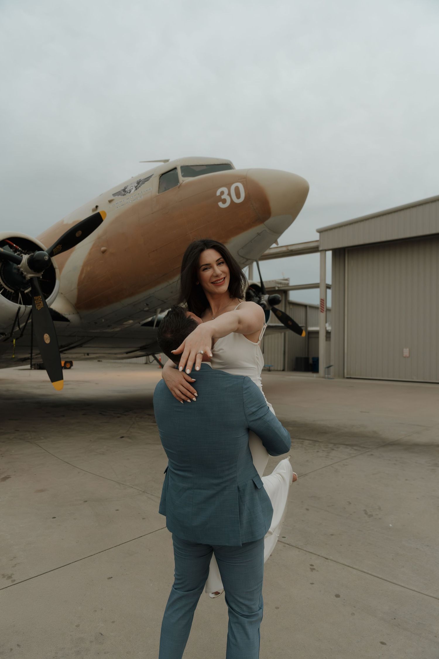 Couple embracing in front of a vintage propeller plane numbered 30 on an airport tarmac, woman in white dress lifted by partner.
