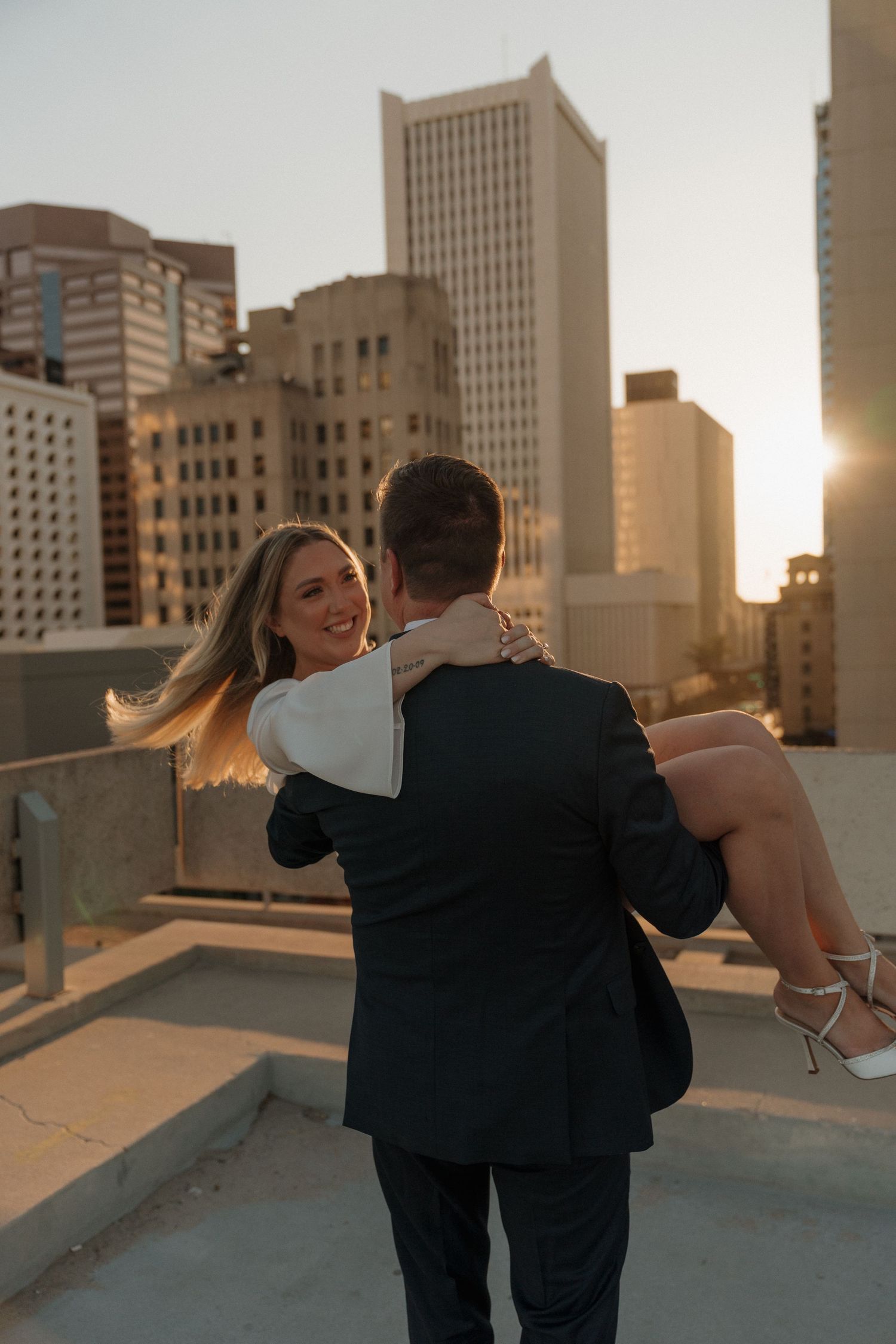 Groom carries smiling bride in white dress on rooftop with city skyline glowing at sunset behind them.