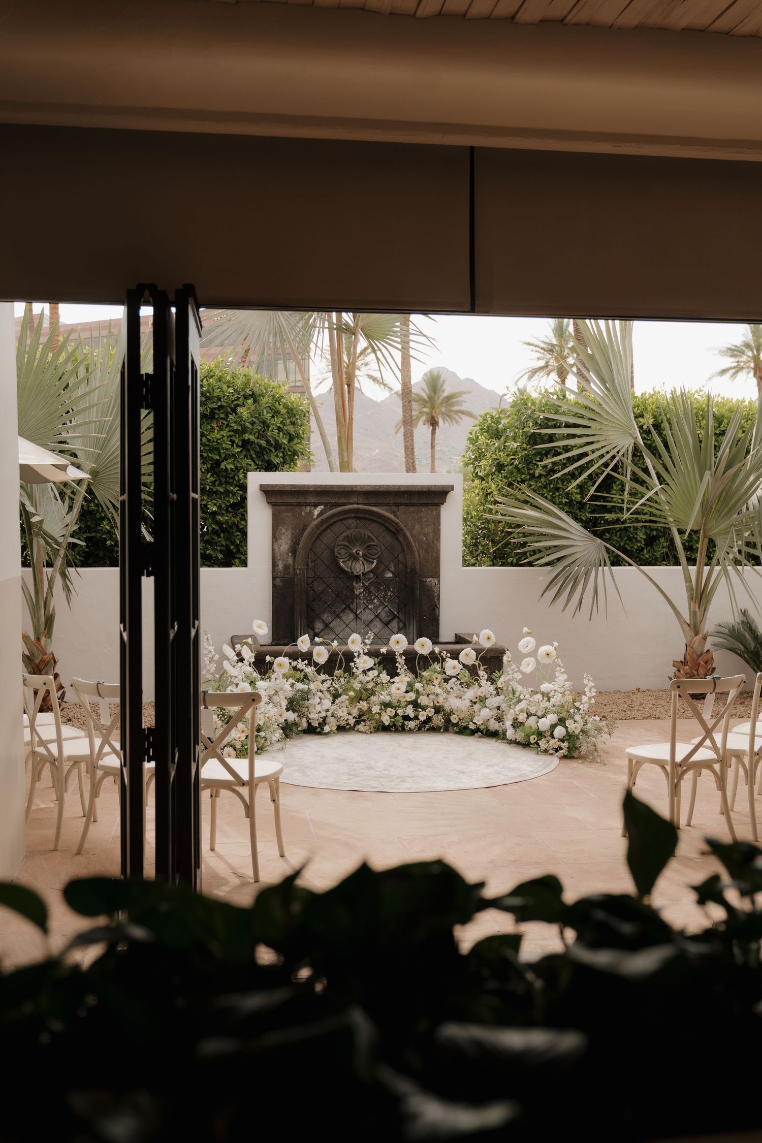 Outdoor wedding ceremony setup with floral arch at stone fountain surrounded by palm trees.