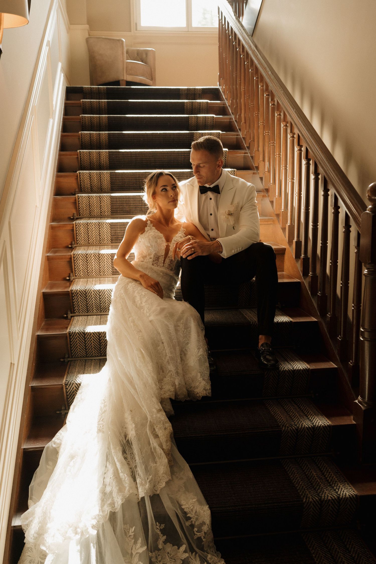 Bride in lace gown and groom in white tuxedo sitting together on wooden staircase indoors.