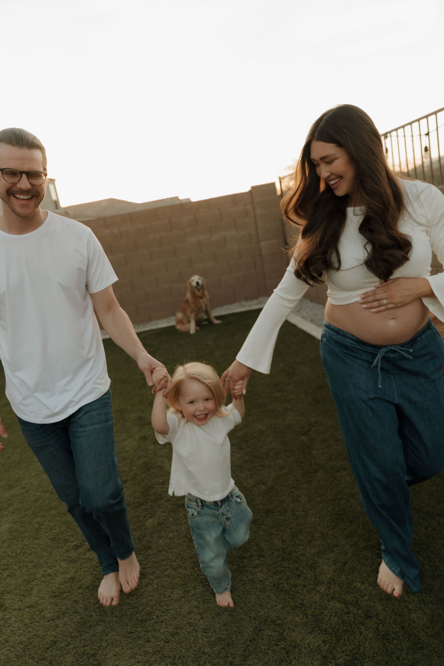 Happy family with toddler and pregnant mother holding hands in backyard with golden retriever.
