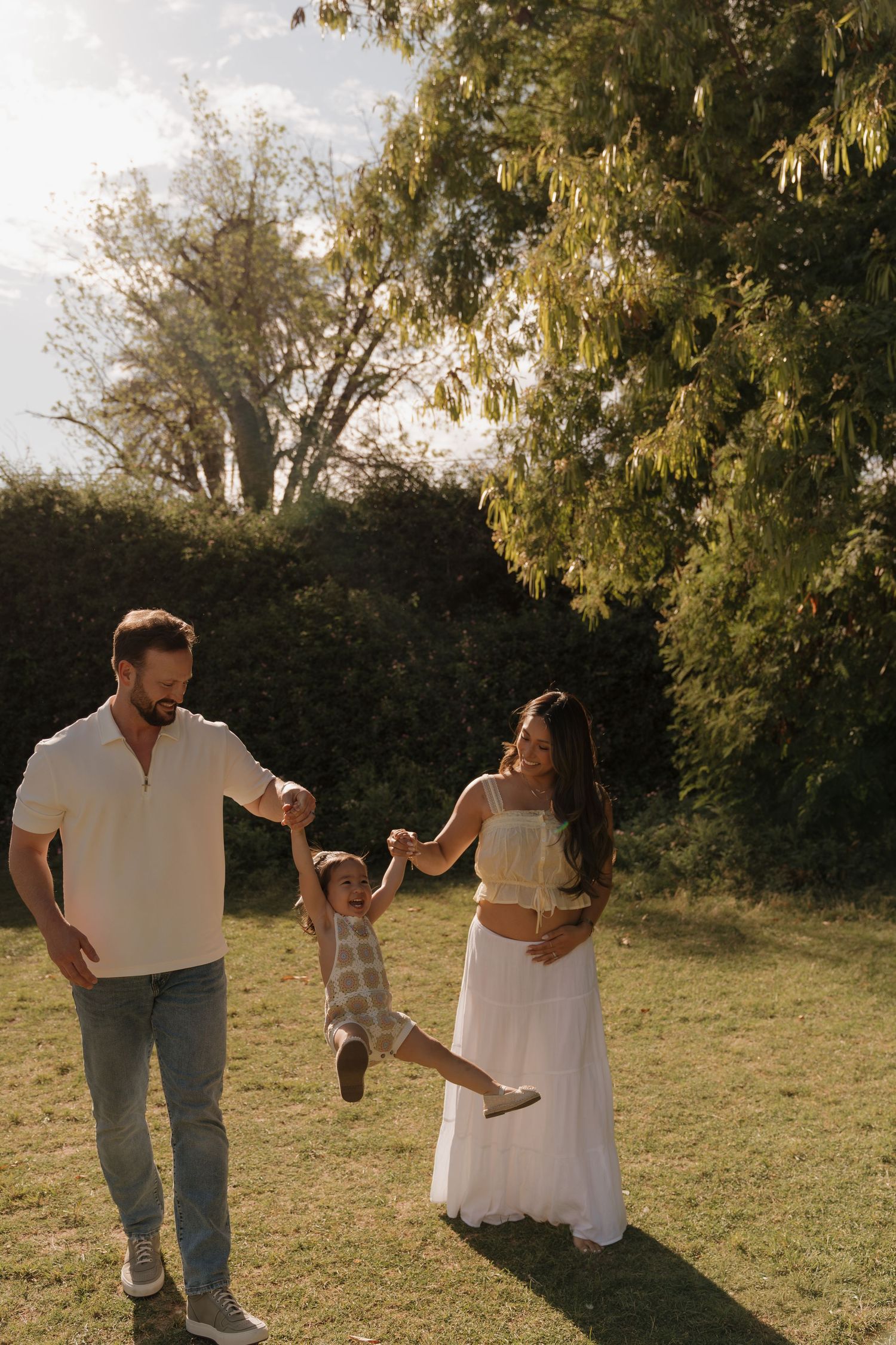 Happy family swinging toddler between parents in sunlit garden with lush green trees behind.