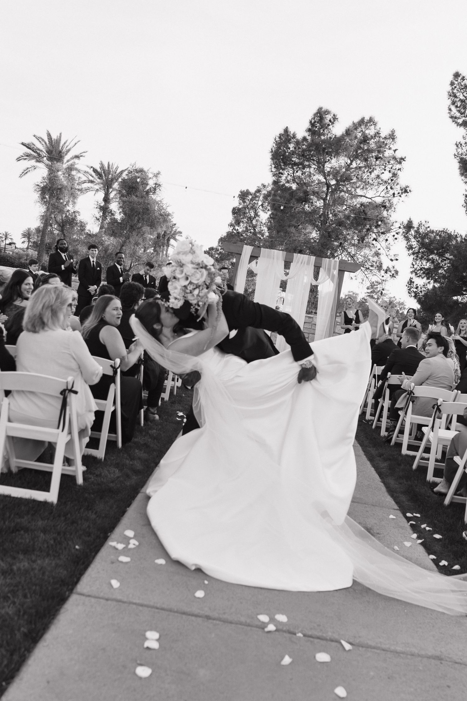 Black and white photo of groom dipping bride at outdoor wedding ceremony aisle.