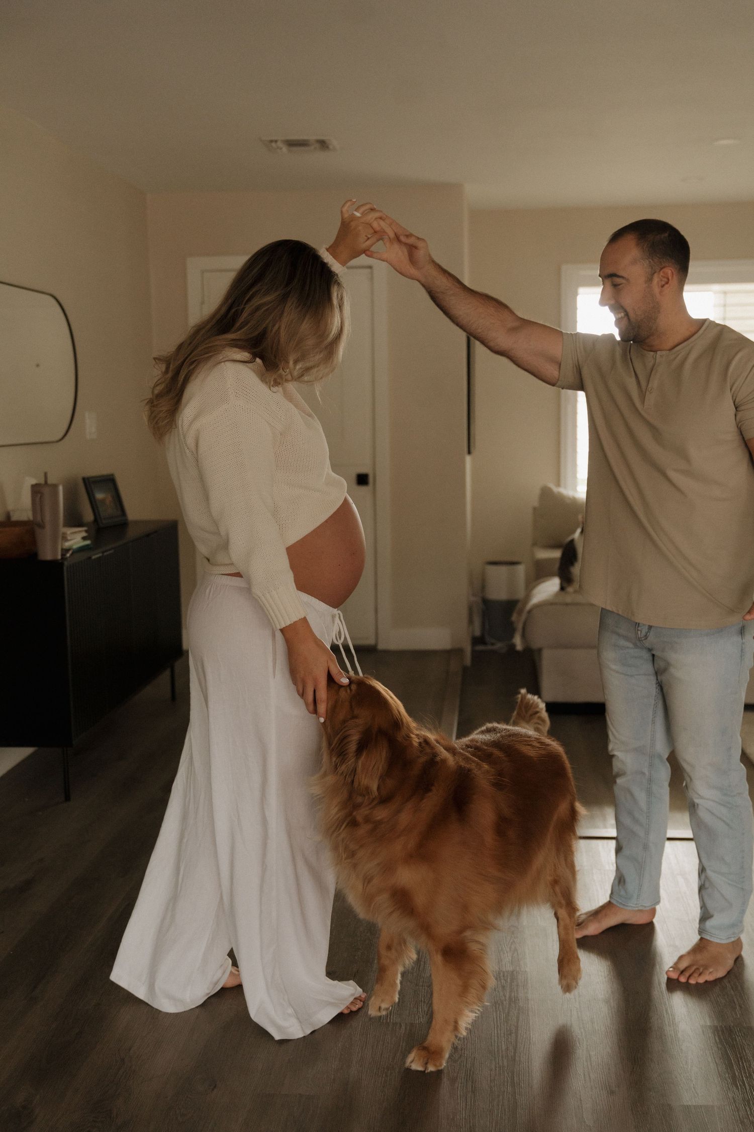 Pregnant woman dances with partner while golden retriever joins in their cozy living room celebration.