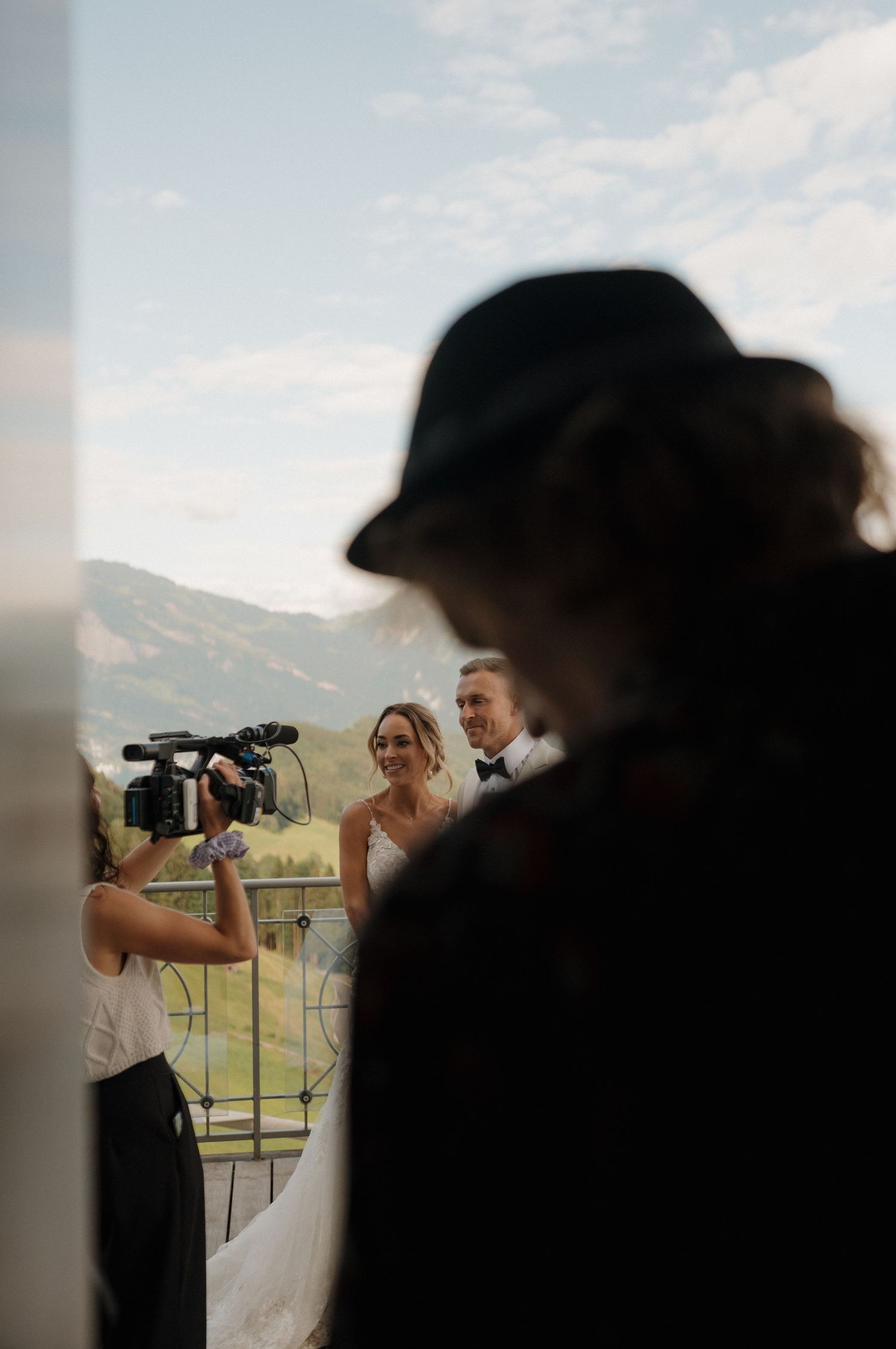 Videographer films smiling bride and groom on a balcony with alpine mountain scenery in the background.
