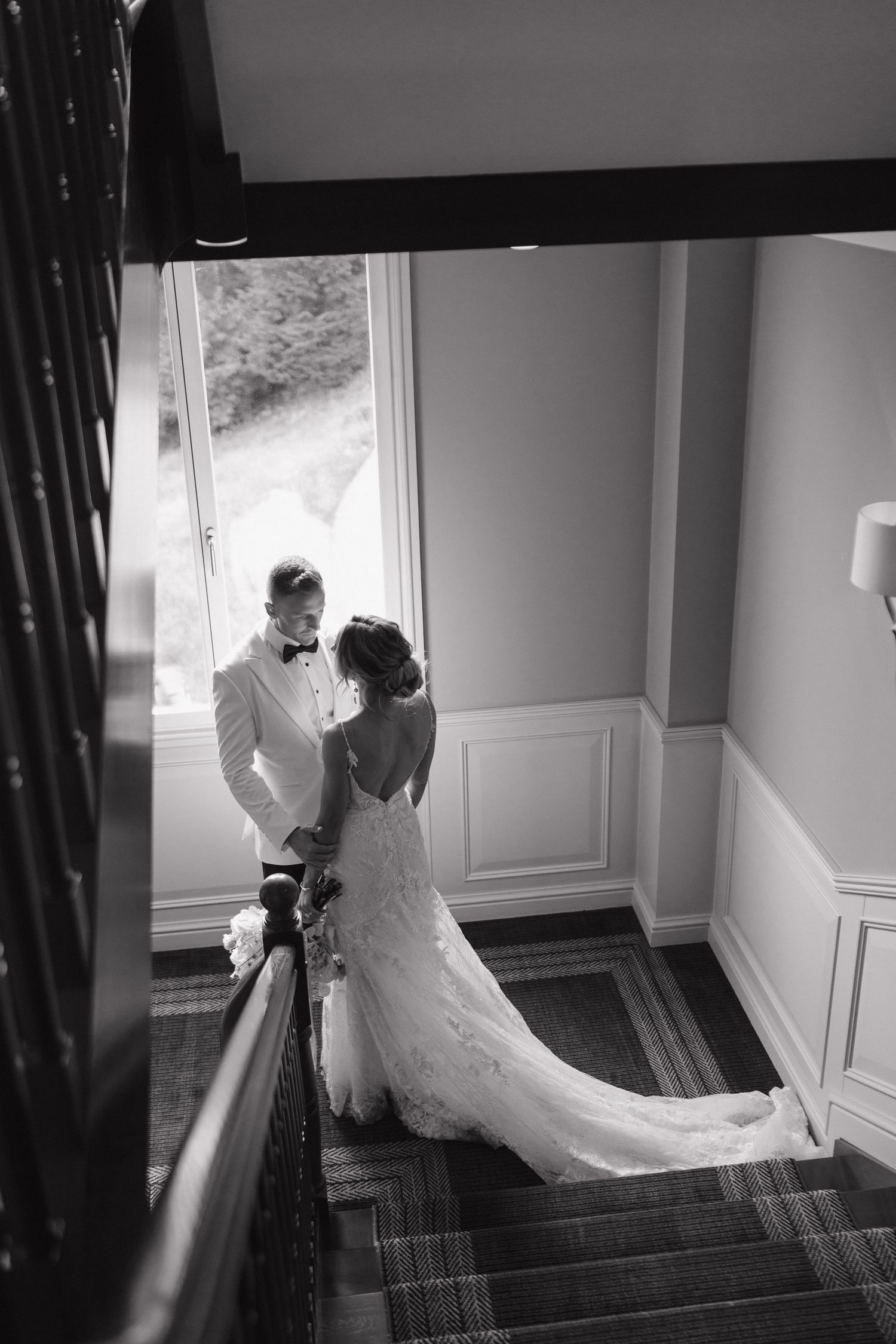 Black and white photo of bride and groom embracing on a staircase near a large window in an elegant venue.
