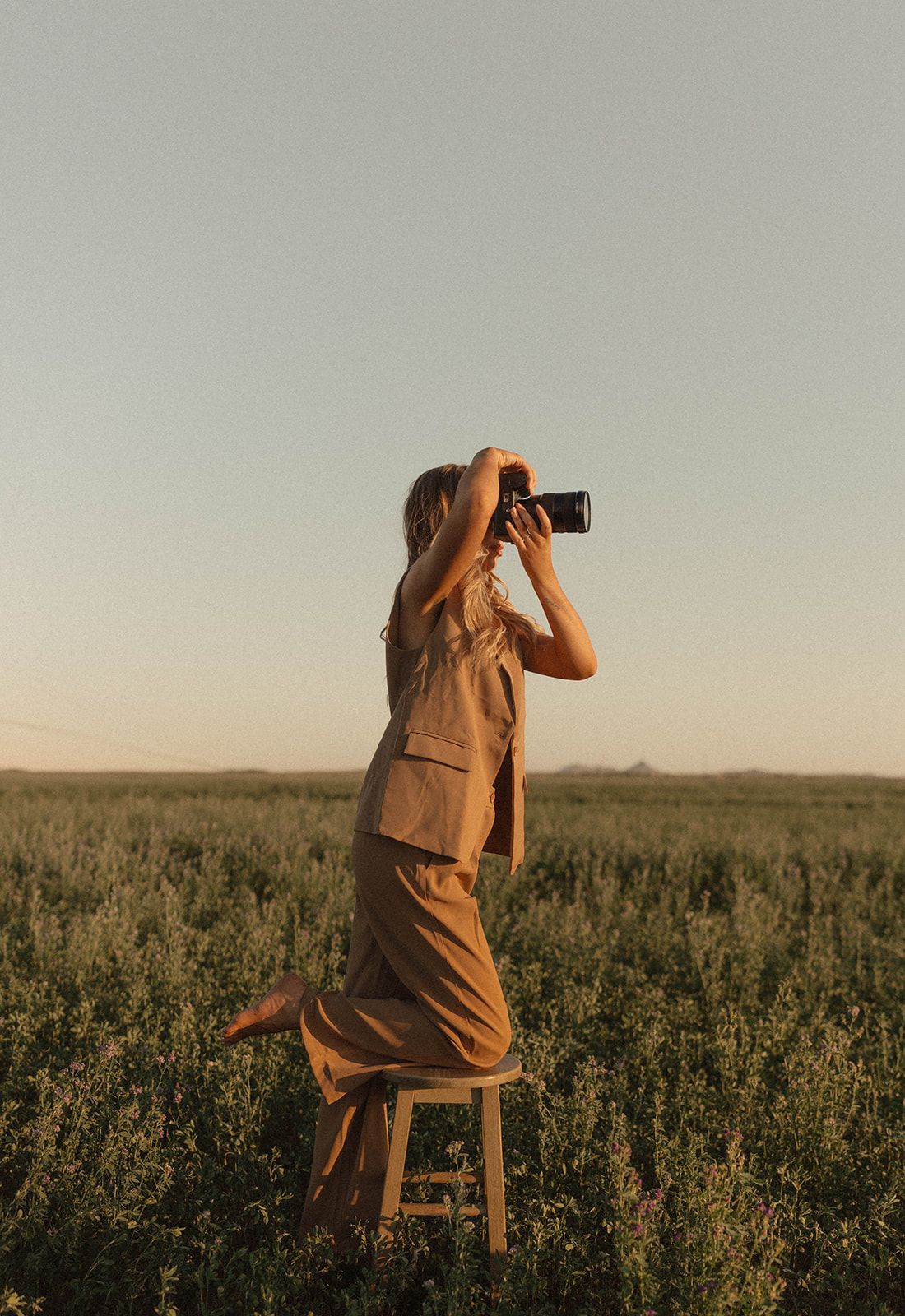 Woman in brown outfit standing on stool in field, photographing landscape during golden hour sunset.