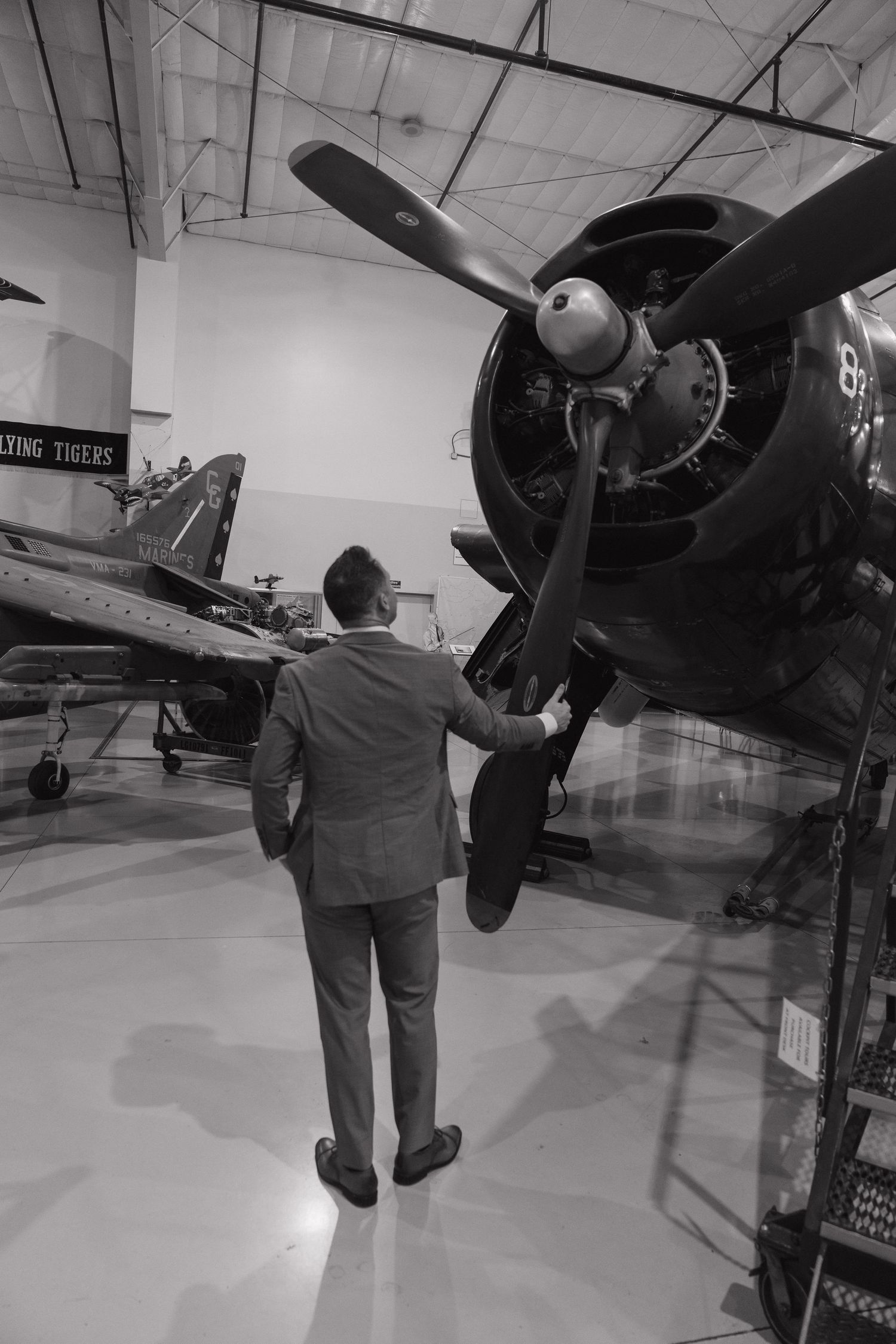 Man in suit examining vintage WWII propeller aircraft engine in Flying Tigers museum hangar, black and white photo.