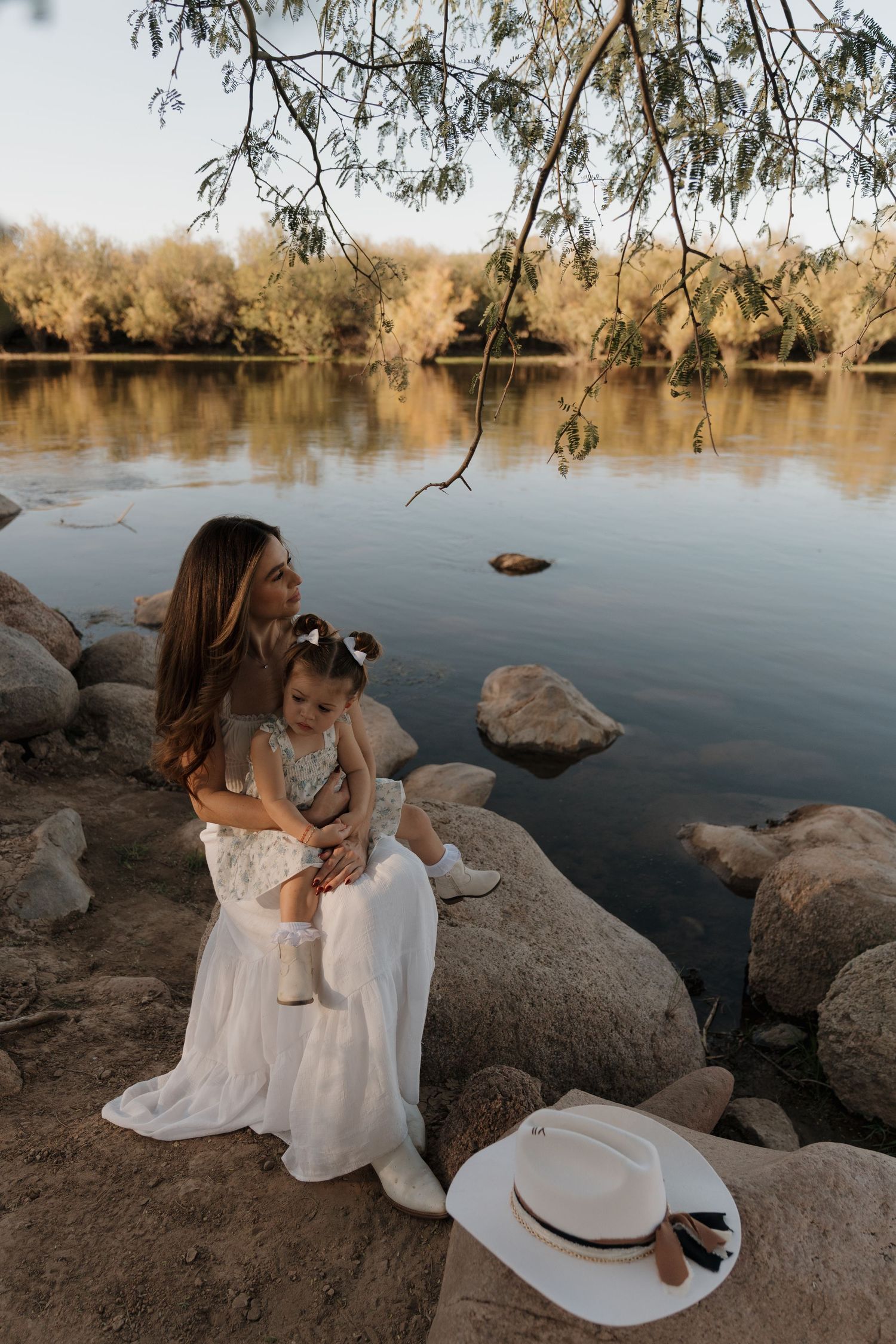 Mother in white dress sitting on rocks by the salt river holding young daughter at sunset.
