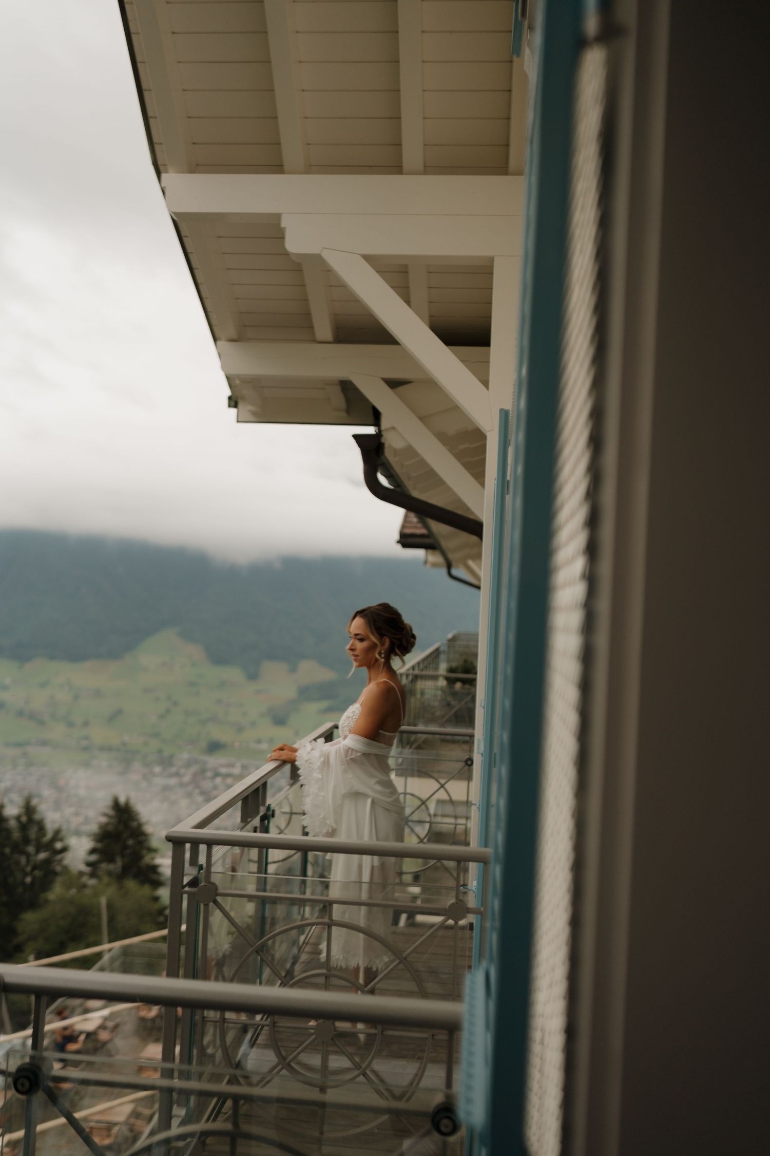Bride in white gown standing on hotel balcony overlooking misty green Alpine mountain landscape.