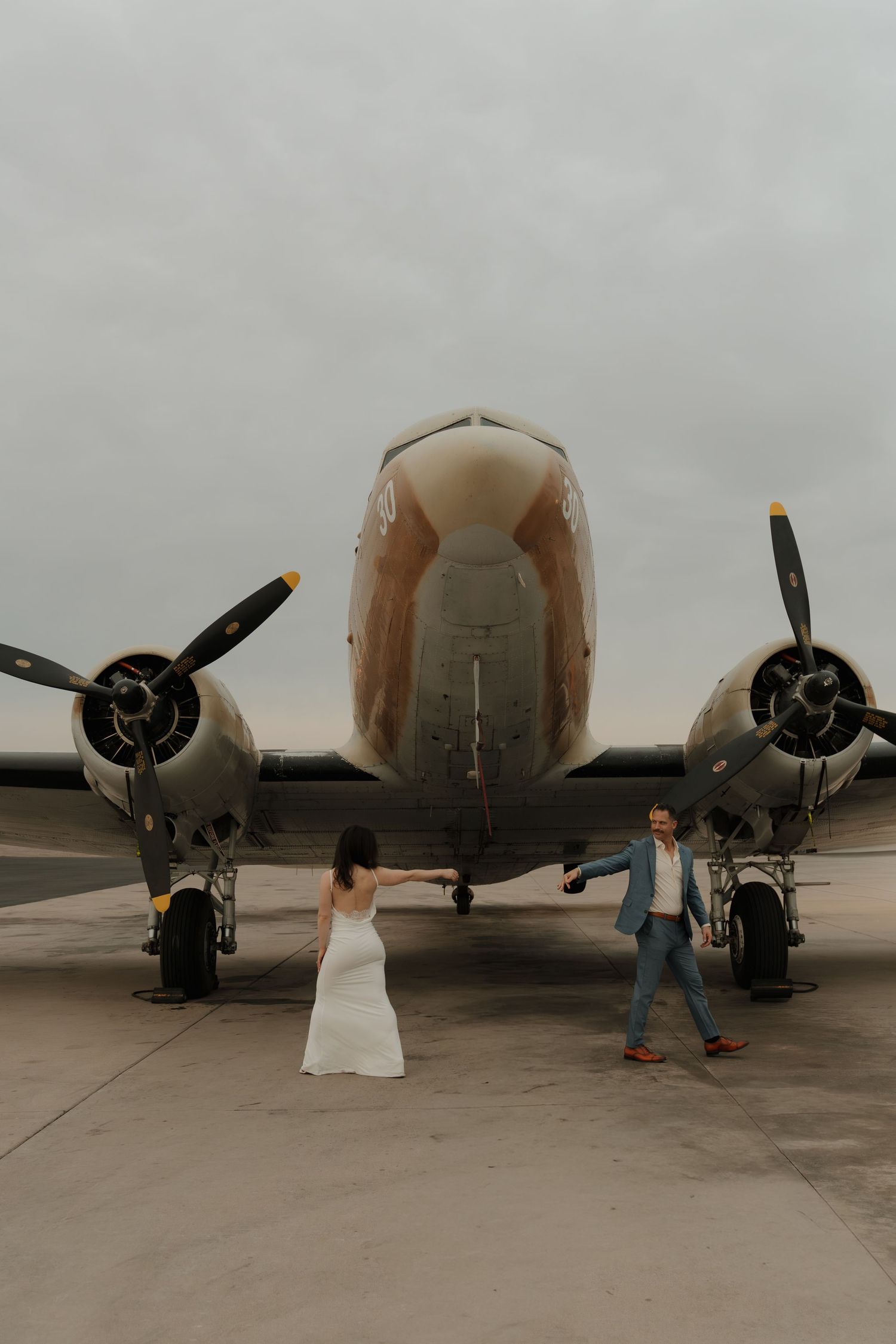 Couple holds hands before twin-engine vintage aircraft on tarmac, woman in white dress, man in blue suit.