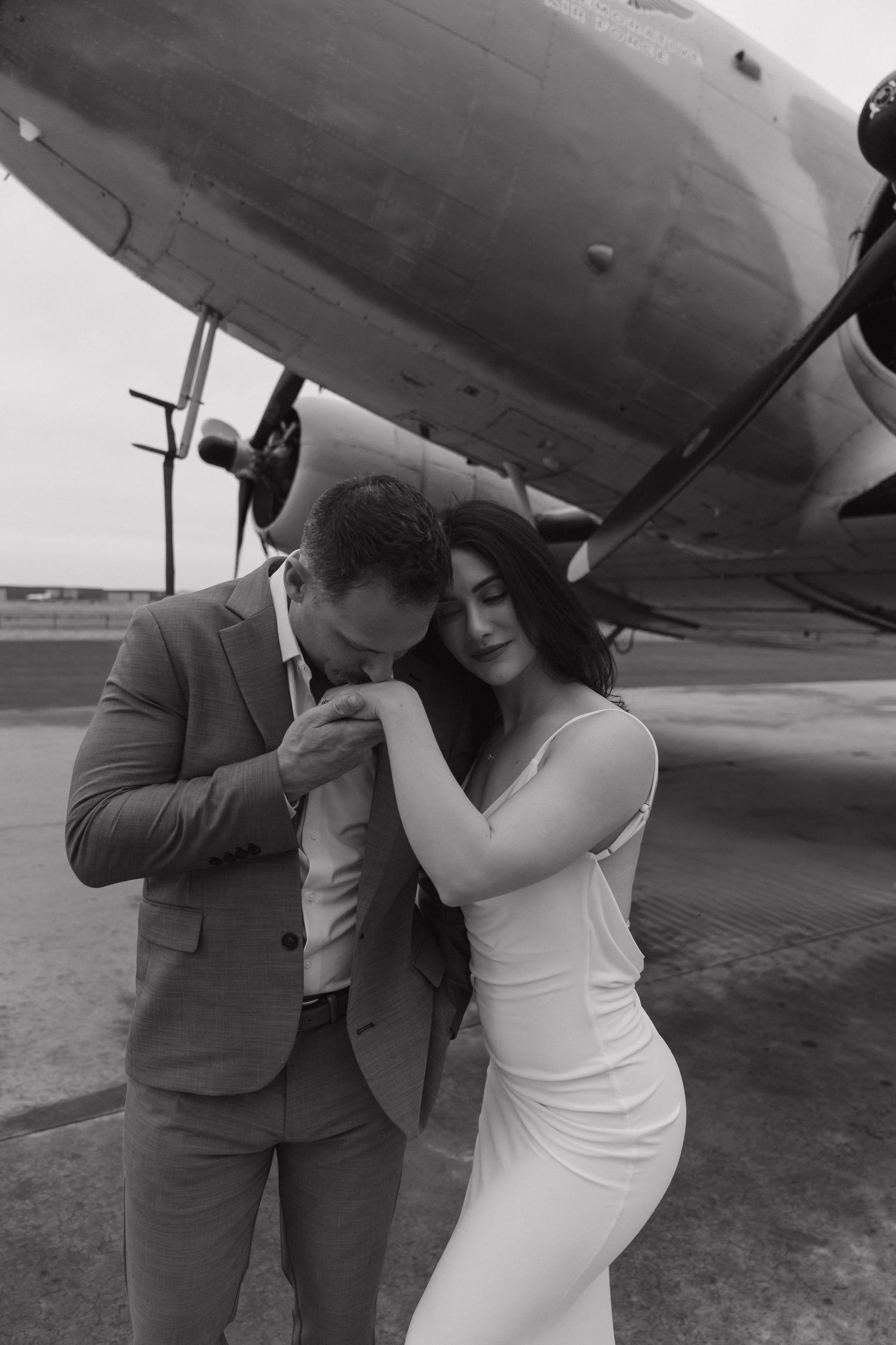 Black and white photo of couple embracing in front of vintage propeller airplane on tarmac.