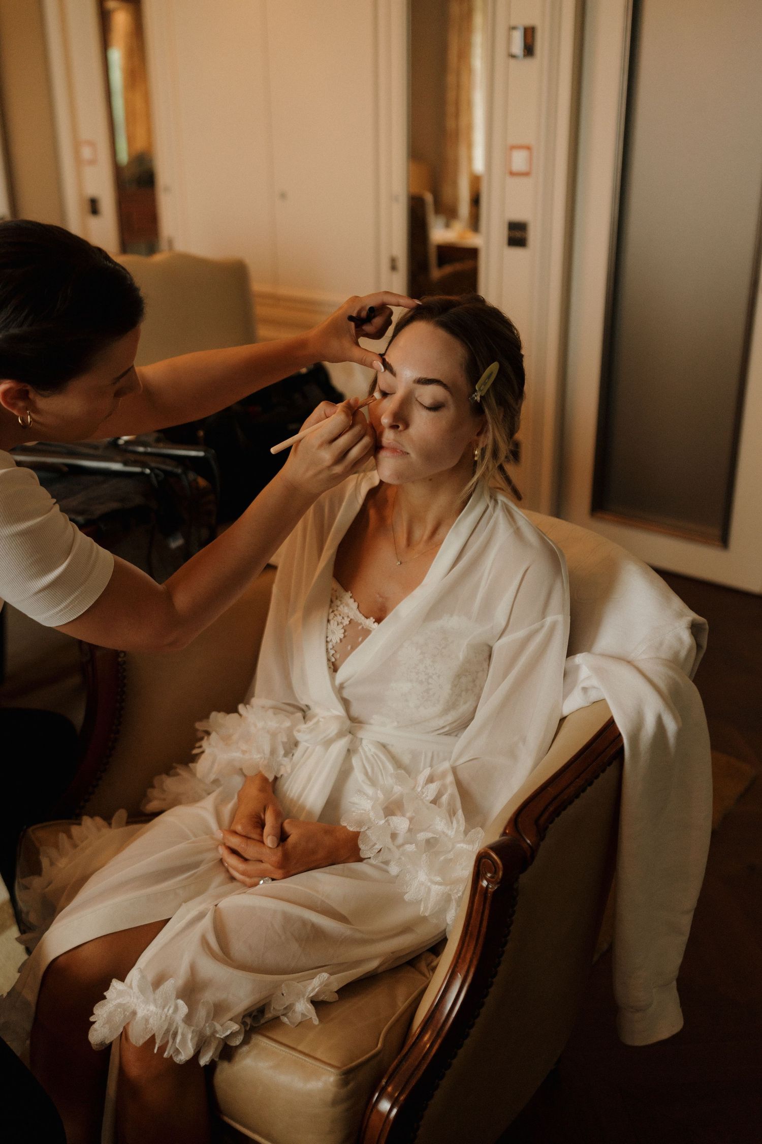 Makeup artist applying eye makeup to bride in white robe seated in elegant chair during wedding prep.