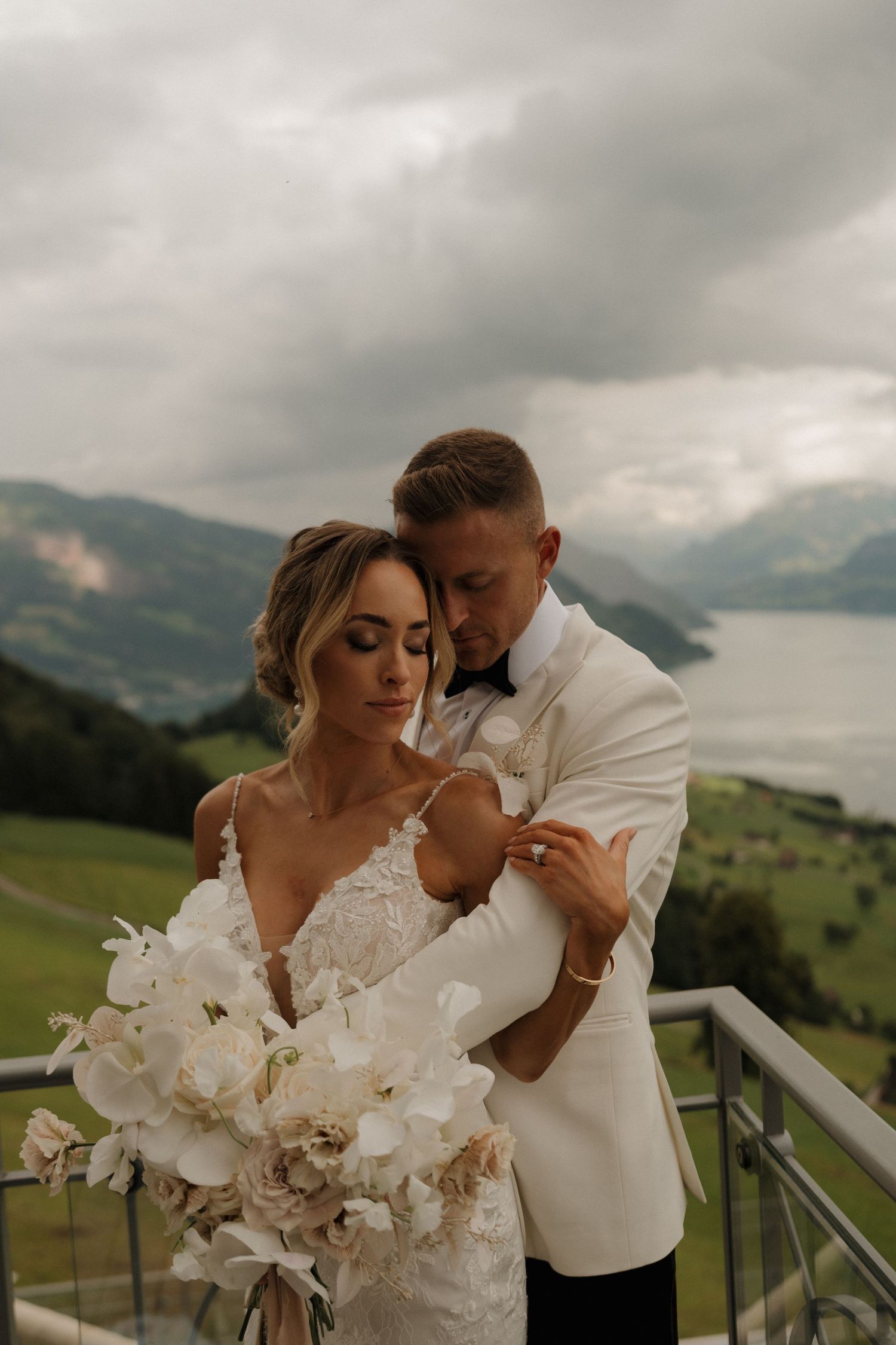 Groom in white tuxedo embracing bride holding white orchid bouquet with mountain lake view.