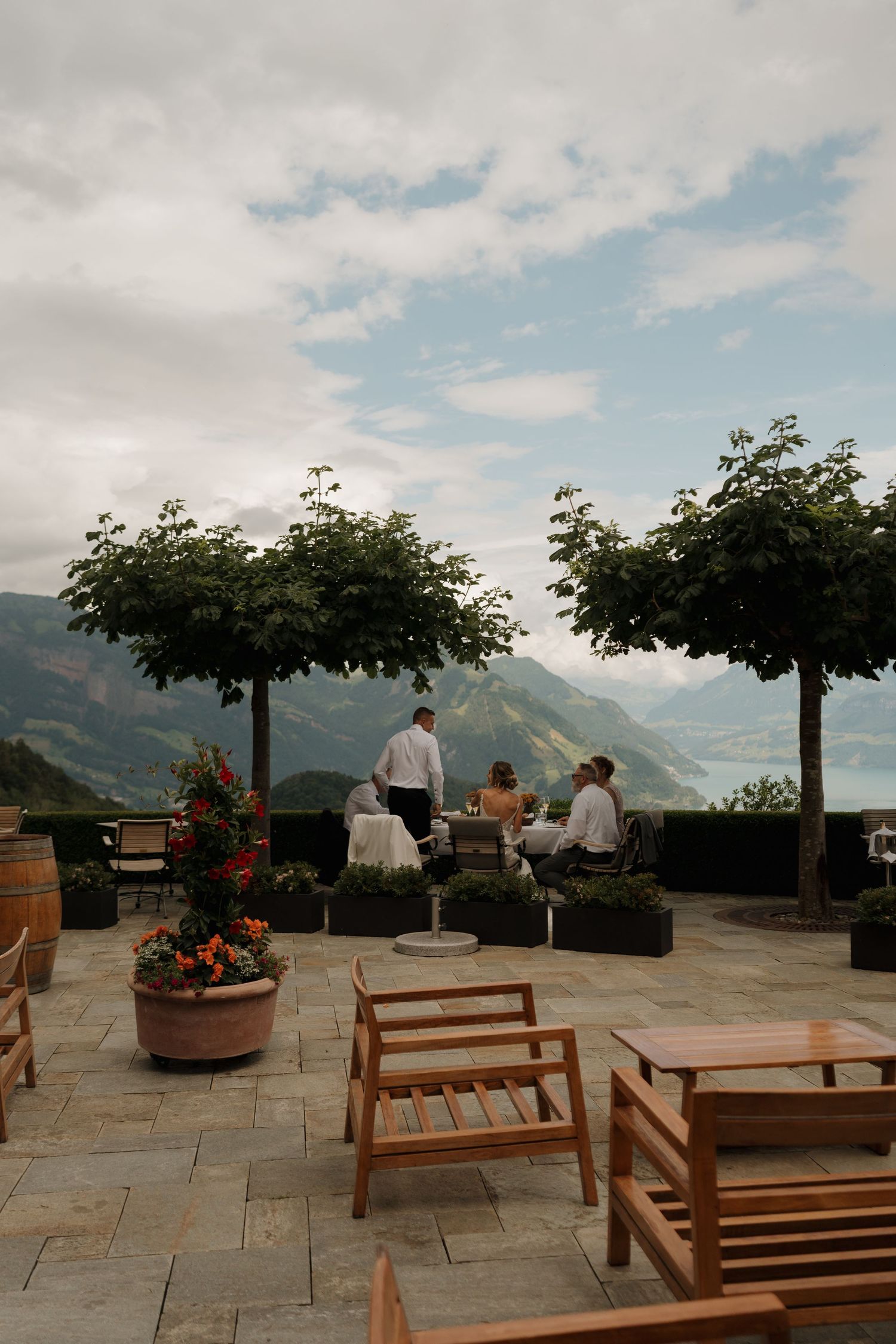 Elegant mountain terrace with wooden chairs, potted trees, and guests enjoying panoramic Alpine views.