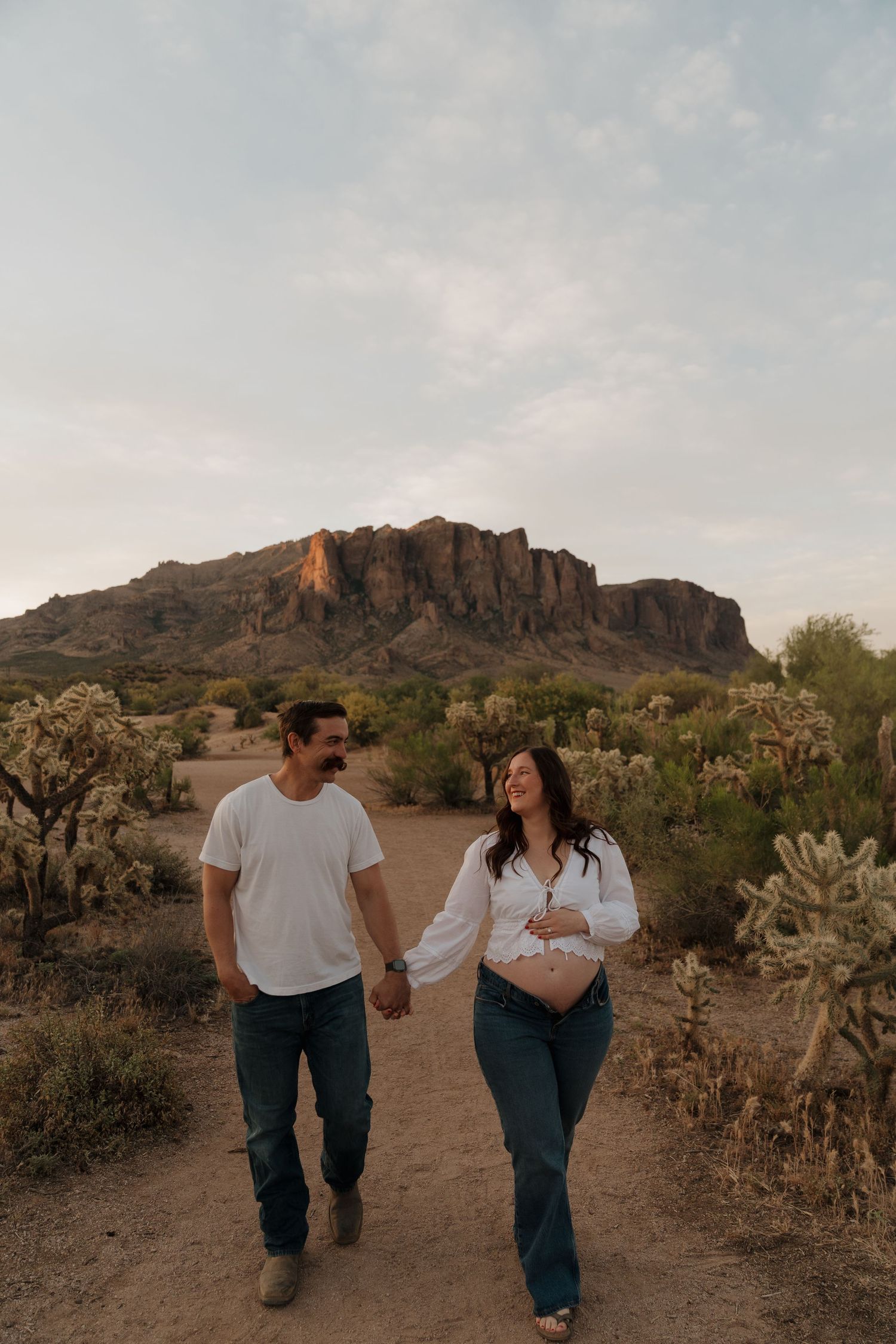 Expecting couple holds hands walking desert path at sunset with rocky mountain backdrop behind them.