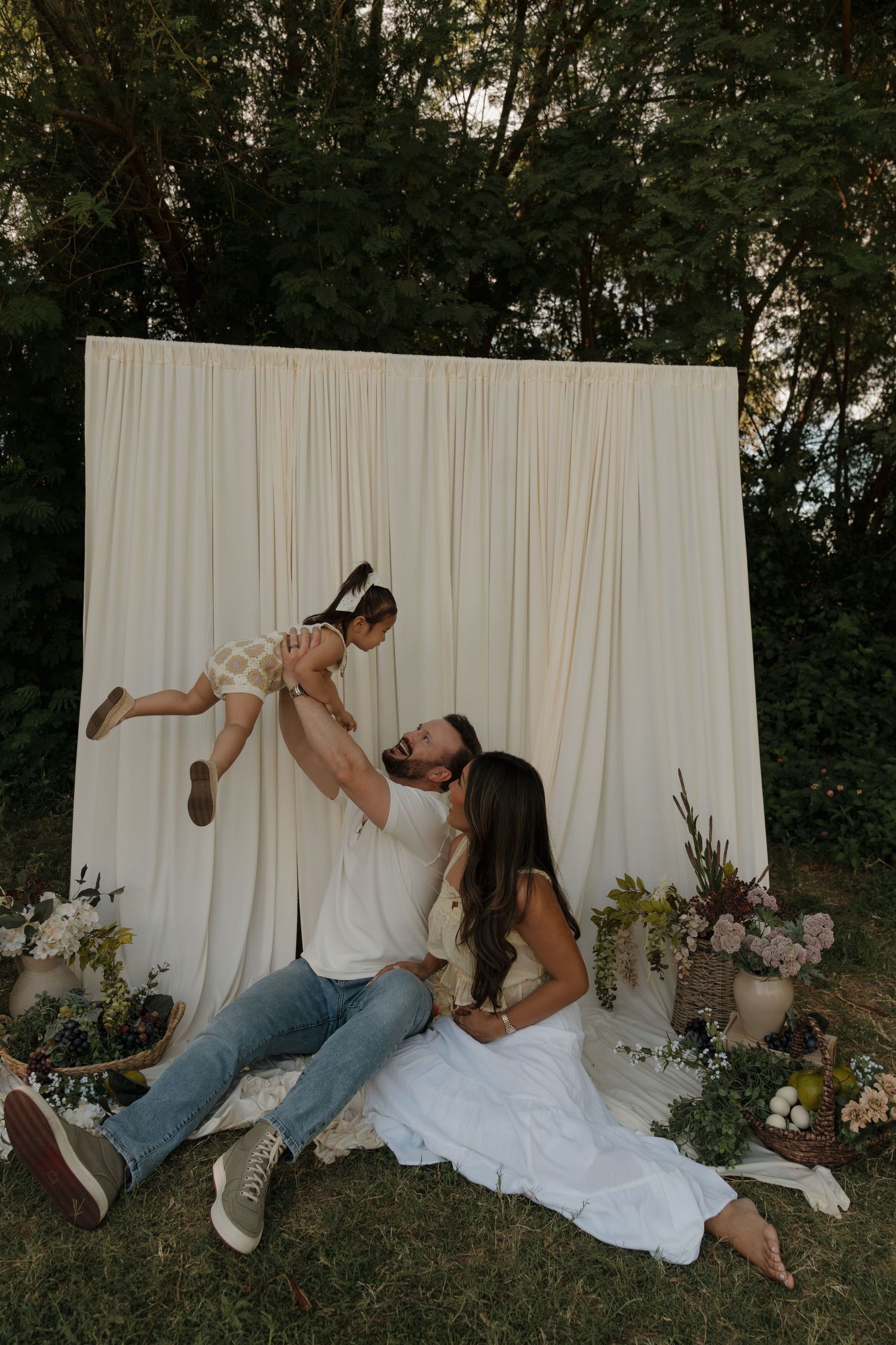 Family sitting before white fabric backdrop with florals as father lifts laughing child overhead.