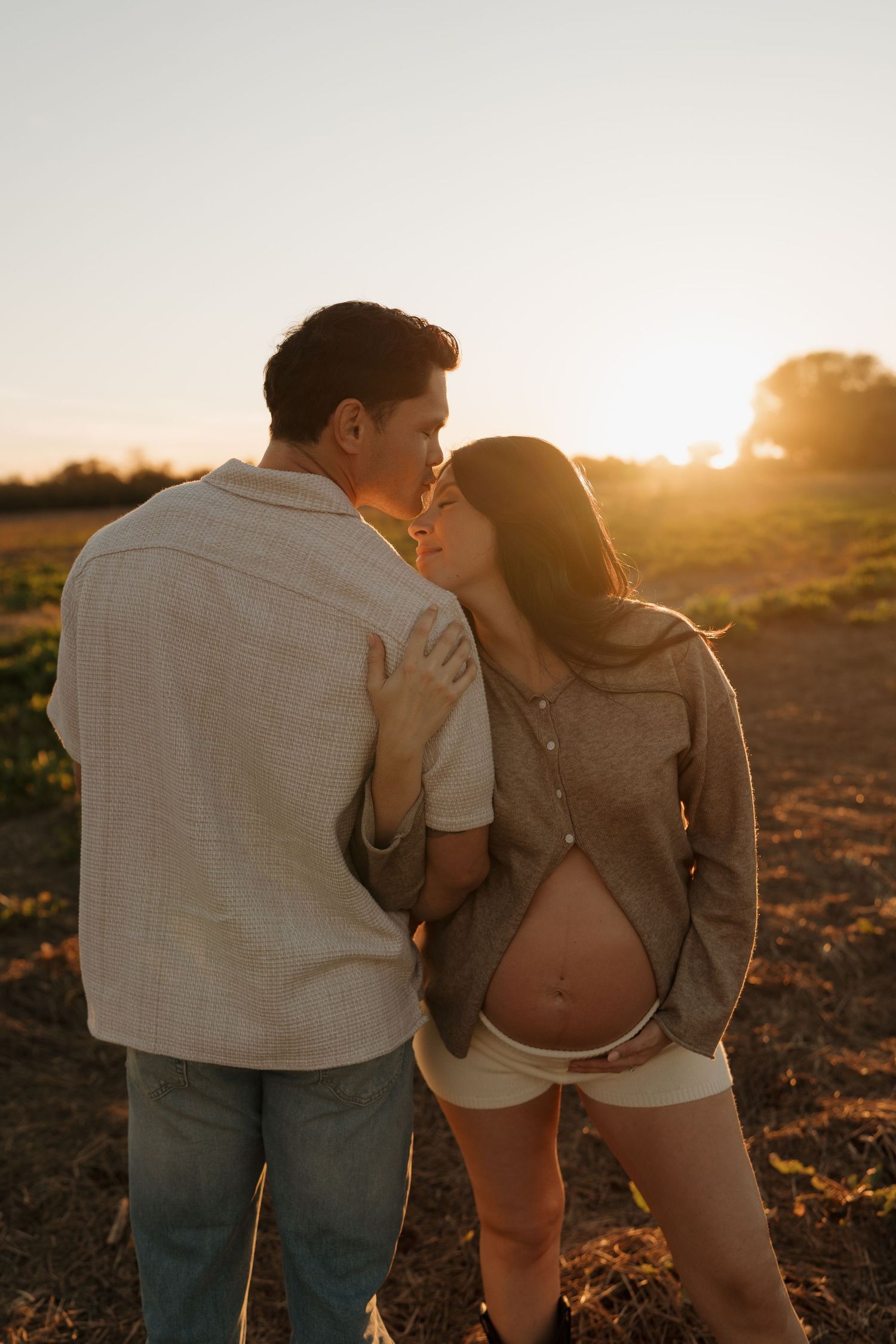 Couple sharing a kiss at sunset in a field, woman showing pregnant belly.