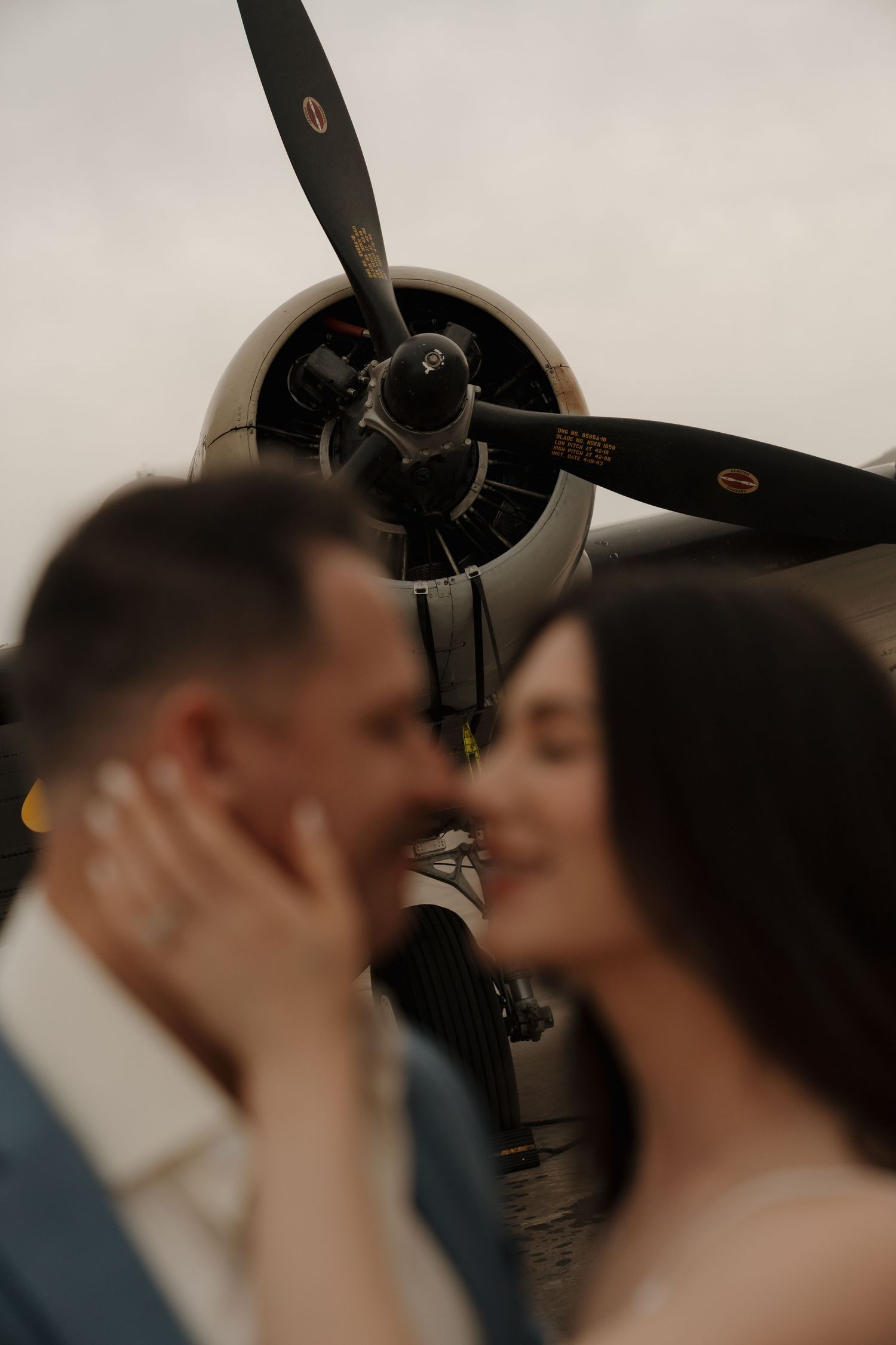 Couple sharing a romantic kiss in front of a vintage propeller airplane, with focus on the aircraft engine.