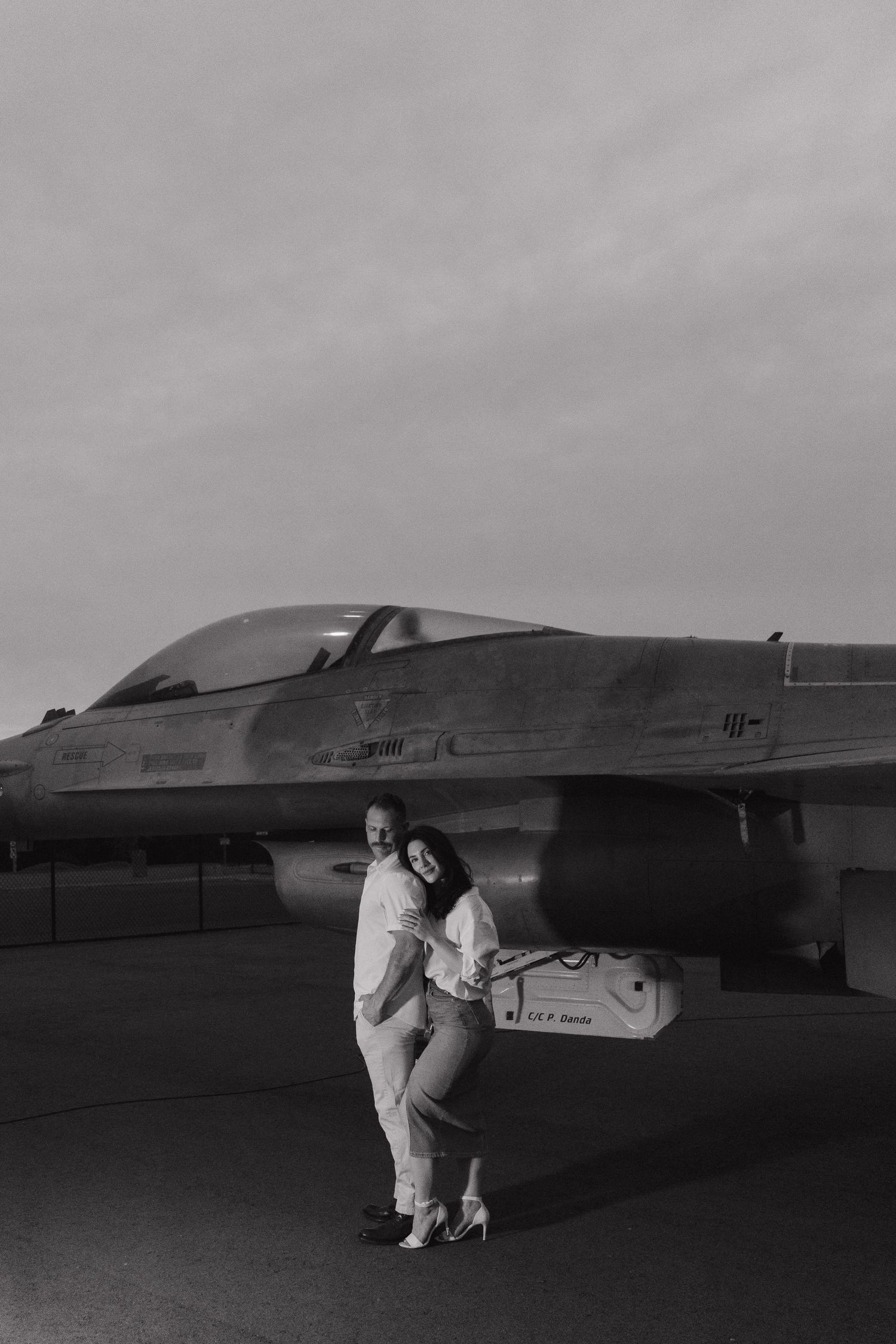 Black and white photo of a couple embracing in front of a military fighter jet on an airfield.