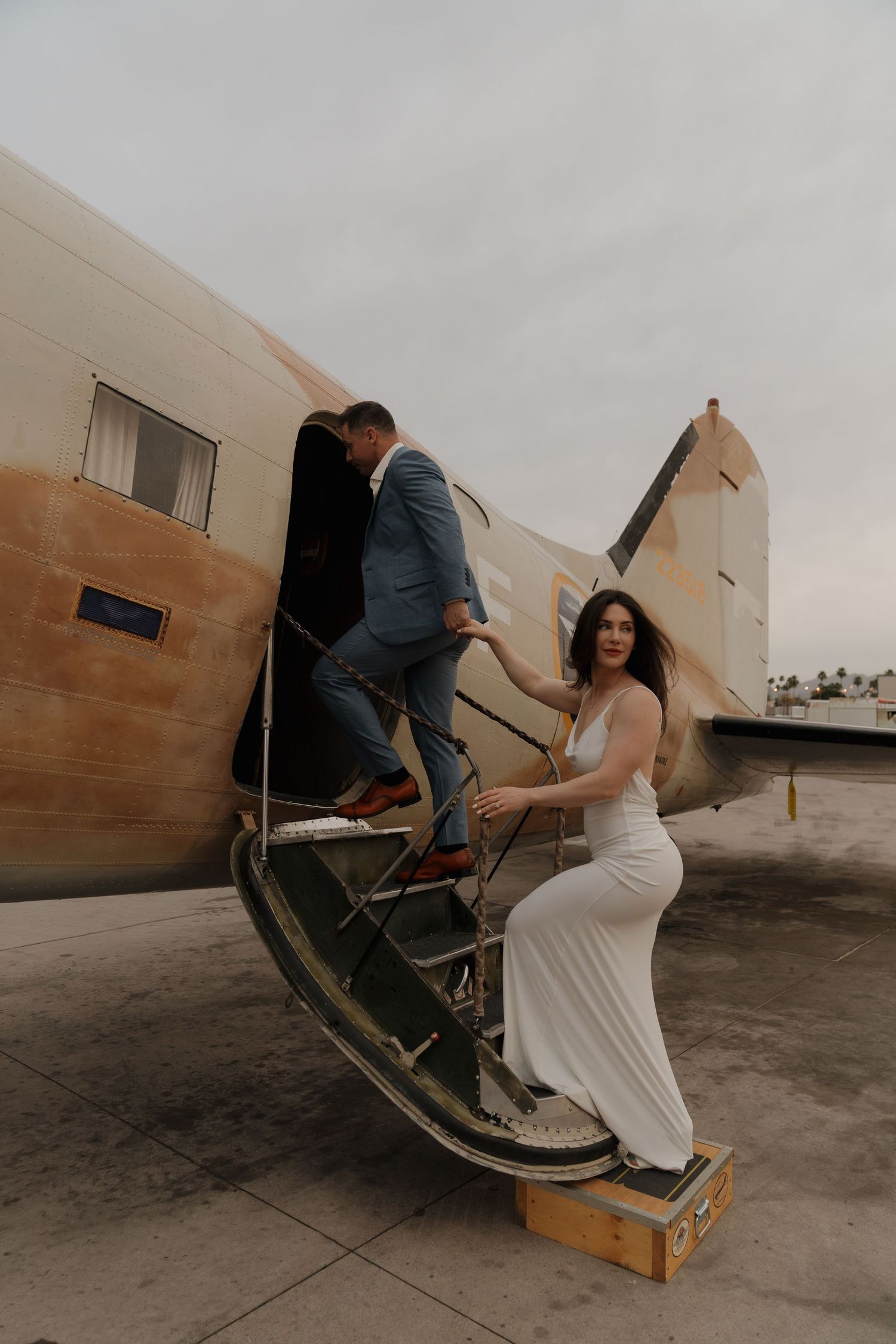 Bride in white gown and groom in blue suit boarding a vintage airplane on their wedding day.