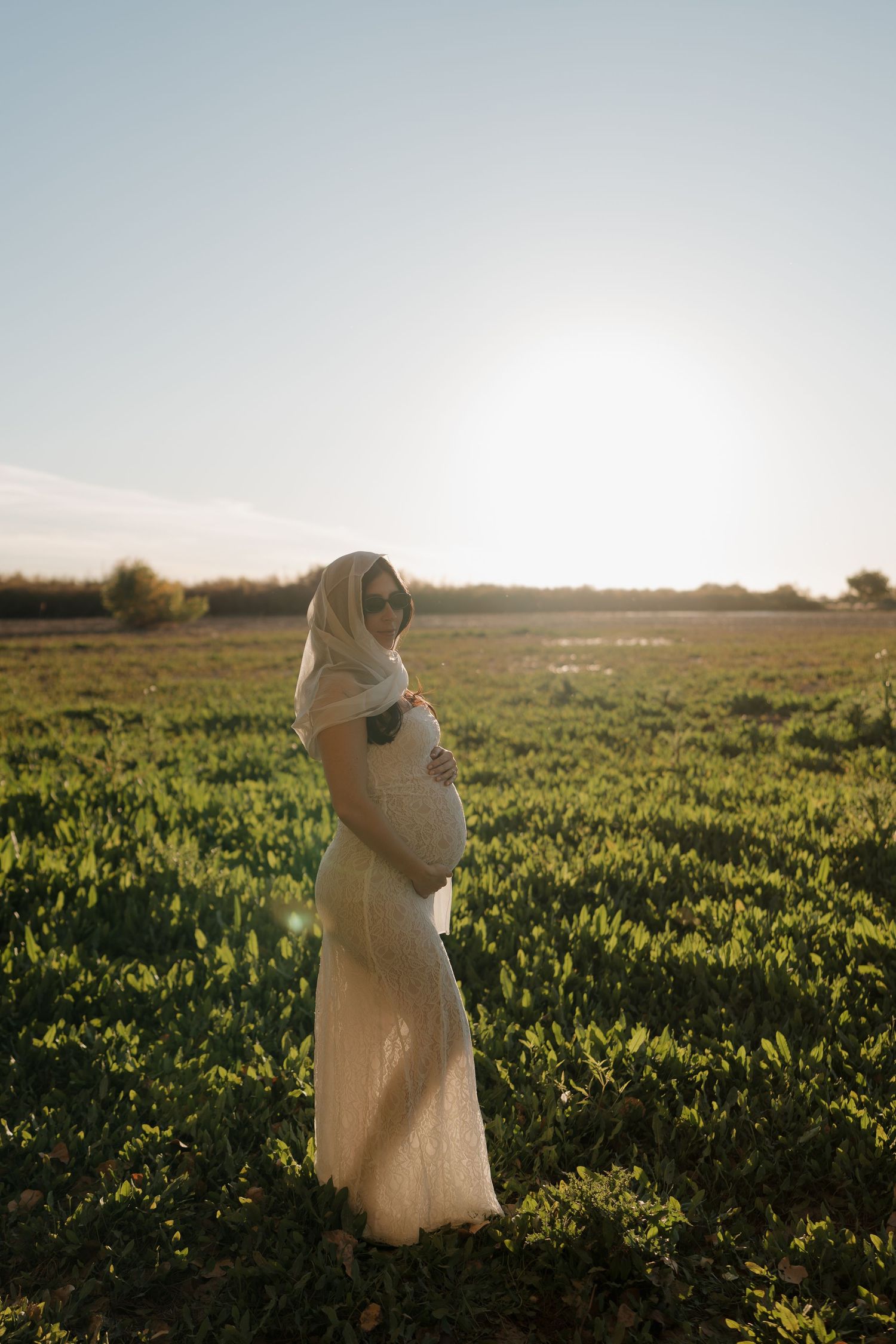 Pregnant woman in white dress standing in green field with sun halo at golden hour.
