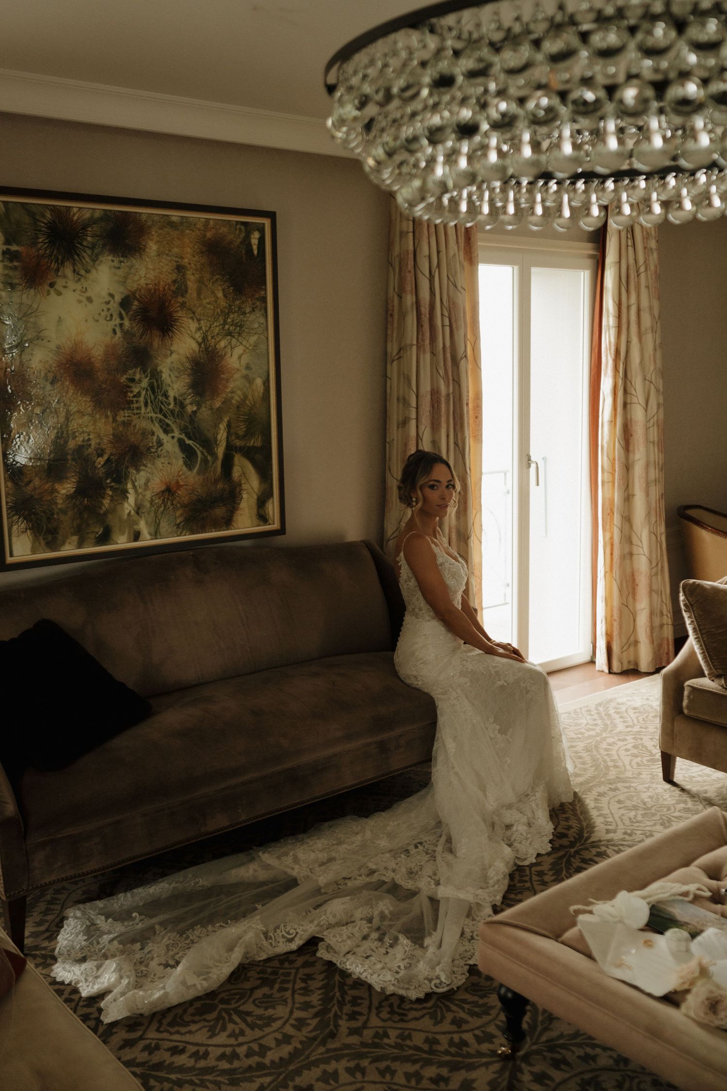 Elegant bride in a lace wedding gown sitting on a velvet sofa beneath a crystal chandelier in a luxury suite.