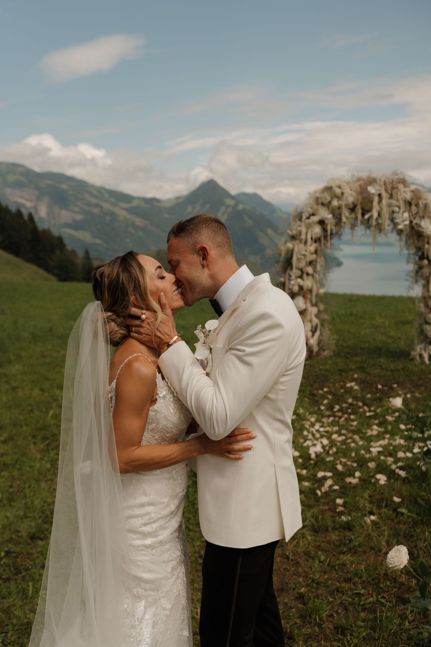 Bride and groom sharing first kiss on a green hillside with a floral arch and lake backdrop.
