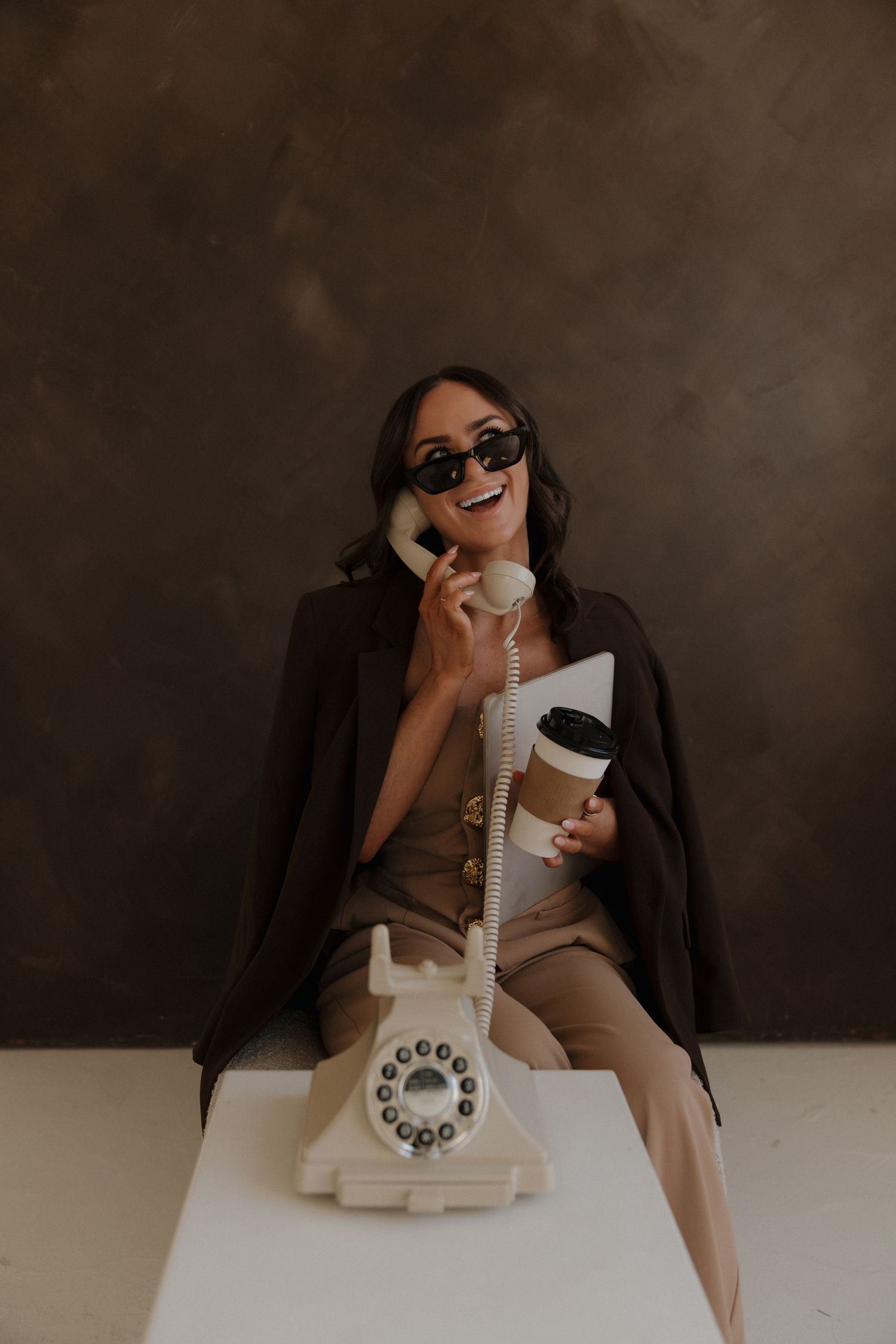Stylish woman in sunglasses talks on vintage rotary phone while holding coffee cup.