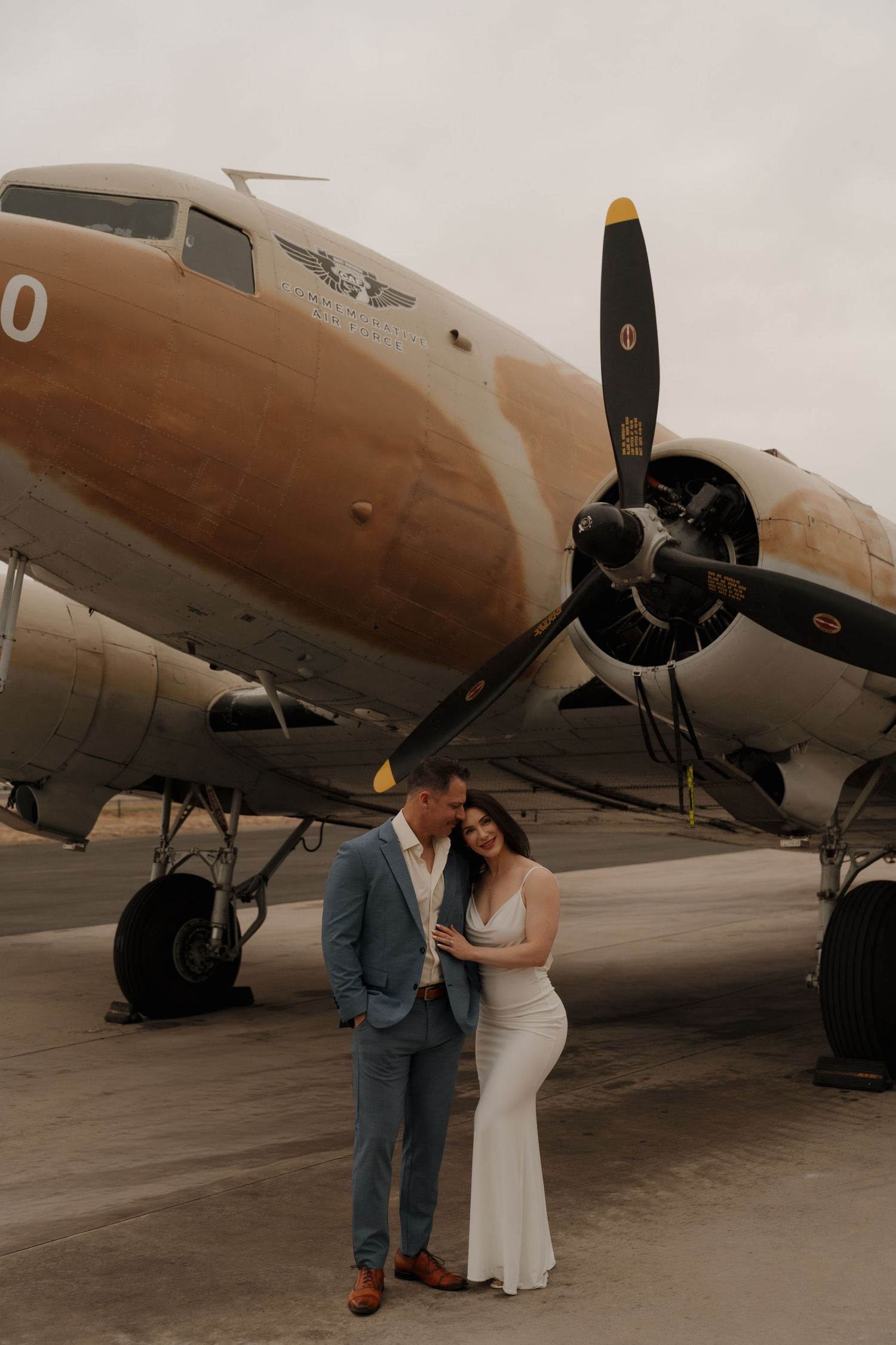 Couple poses romantically in front of a vintage propeller airplane on a tarmac, dressed in wedding attire.
