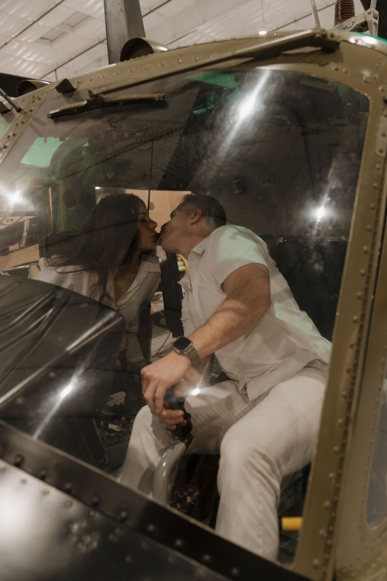 Couple sharing a romantic kiss inside a helicopter cockpit in a hangar, man wearing white outfit holding controls.