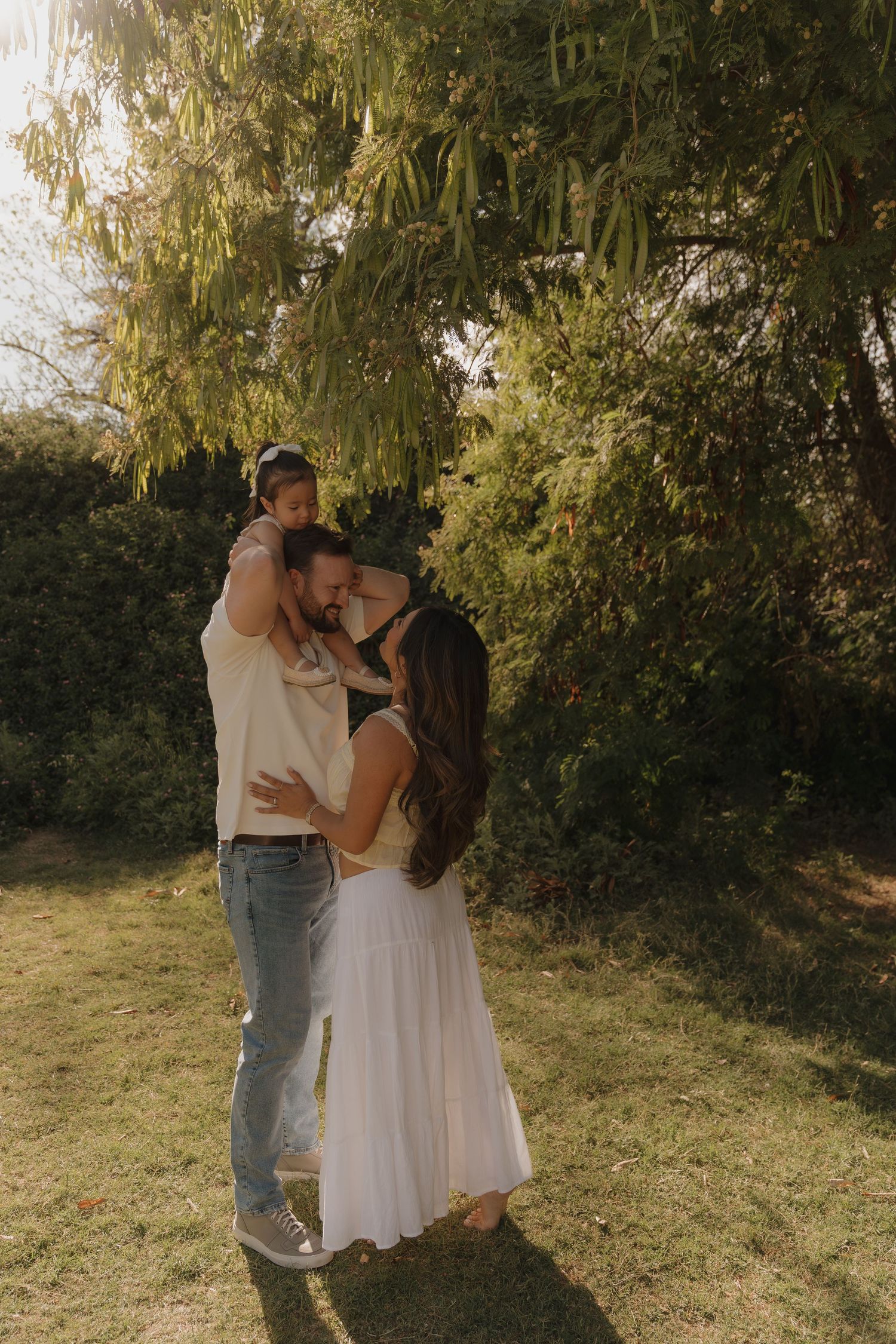 Family of three embracing under weeping willow tree in warm golden afternoon sunlight outdoors.