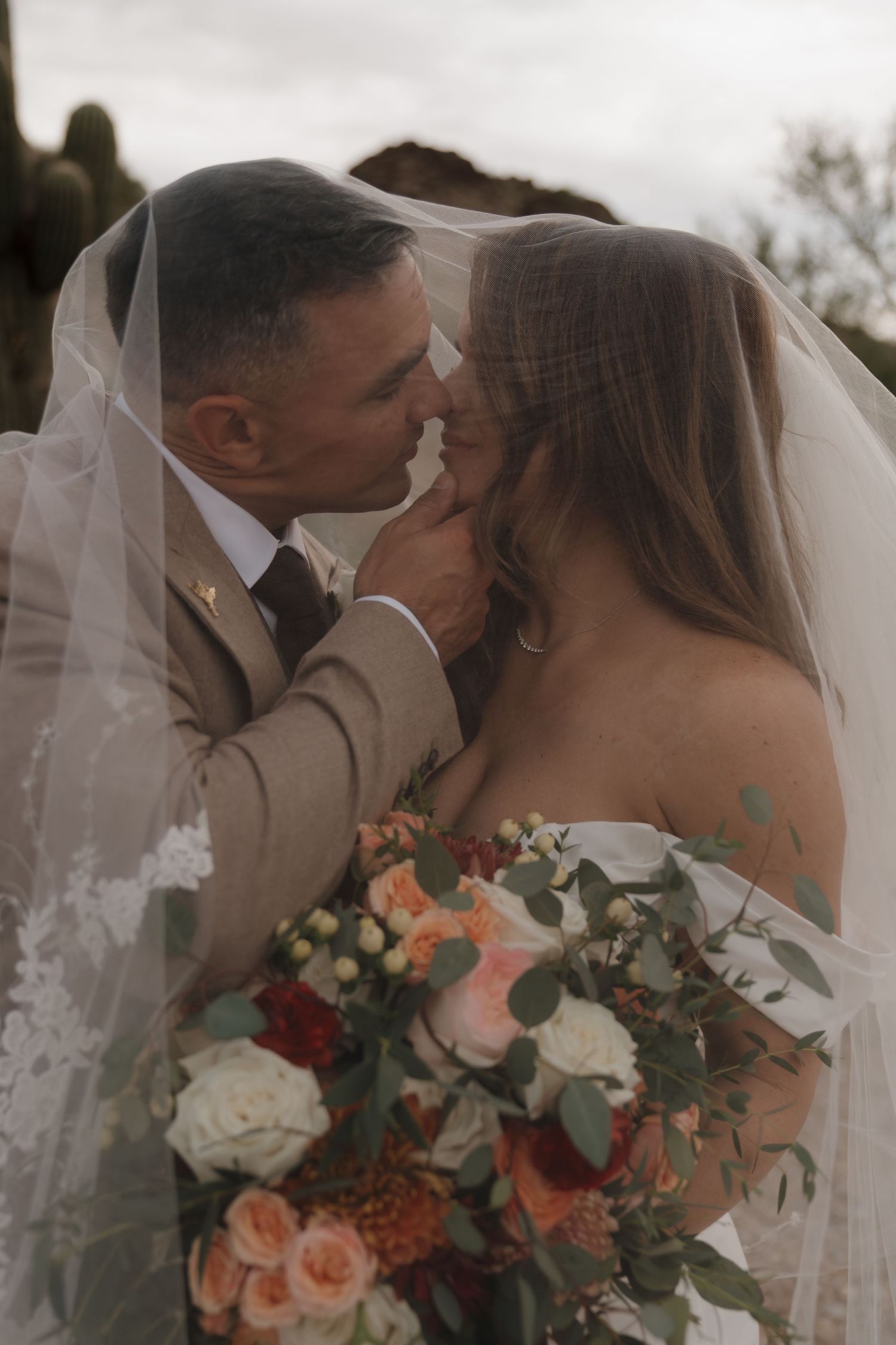 Bride and groom share intimate moment under veil with colorful floral bouquet in desert wedding setting.