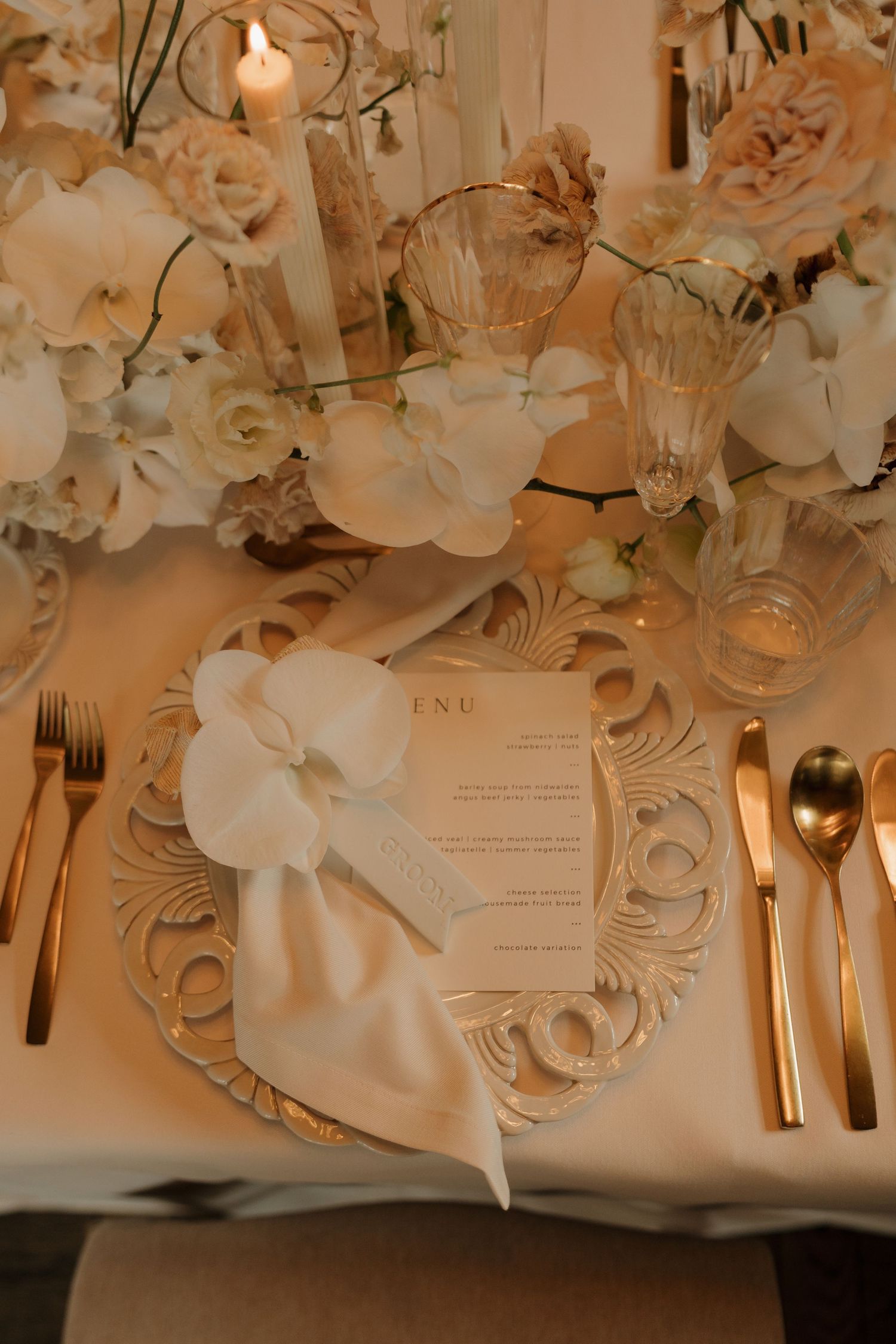 Close-up of wedding table setting featuring white flowers, gold flatware, crystal glasses, and menu card.