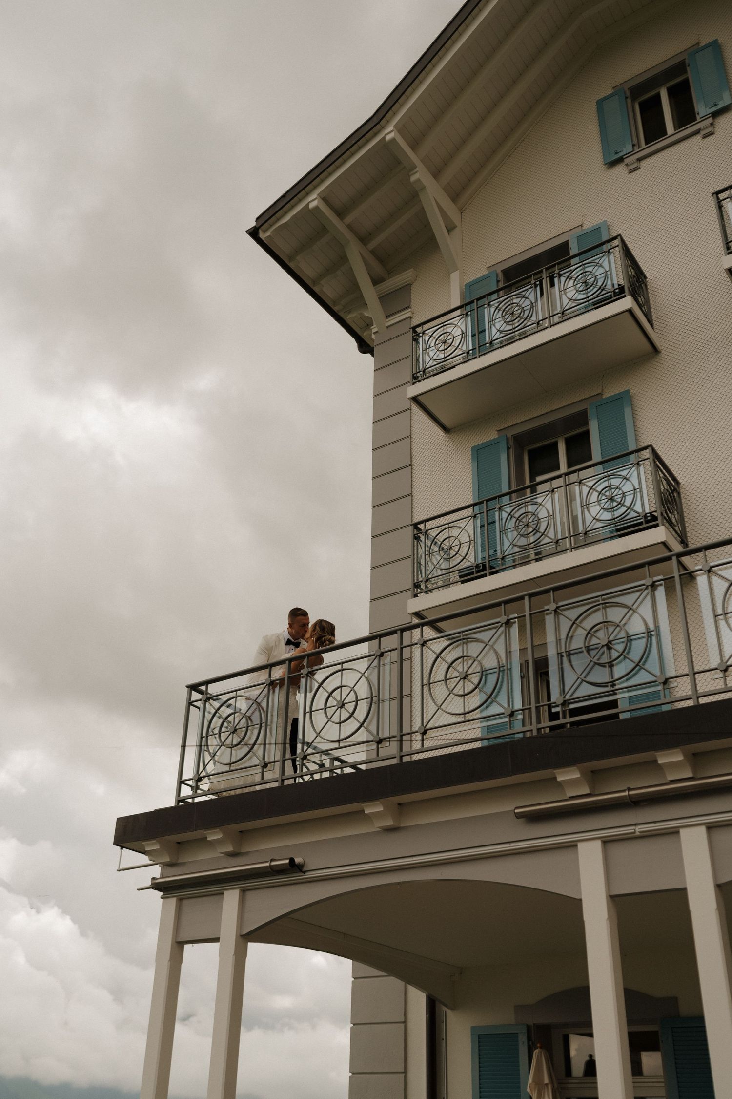 Bride and groom sharing intimate moment on balcony of elegant white building with blue shutters.