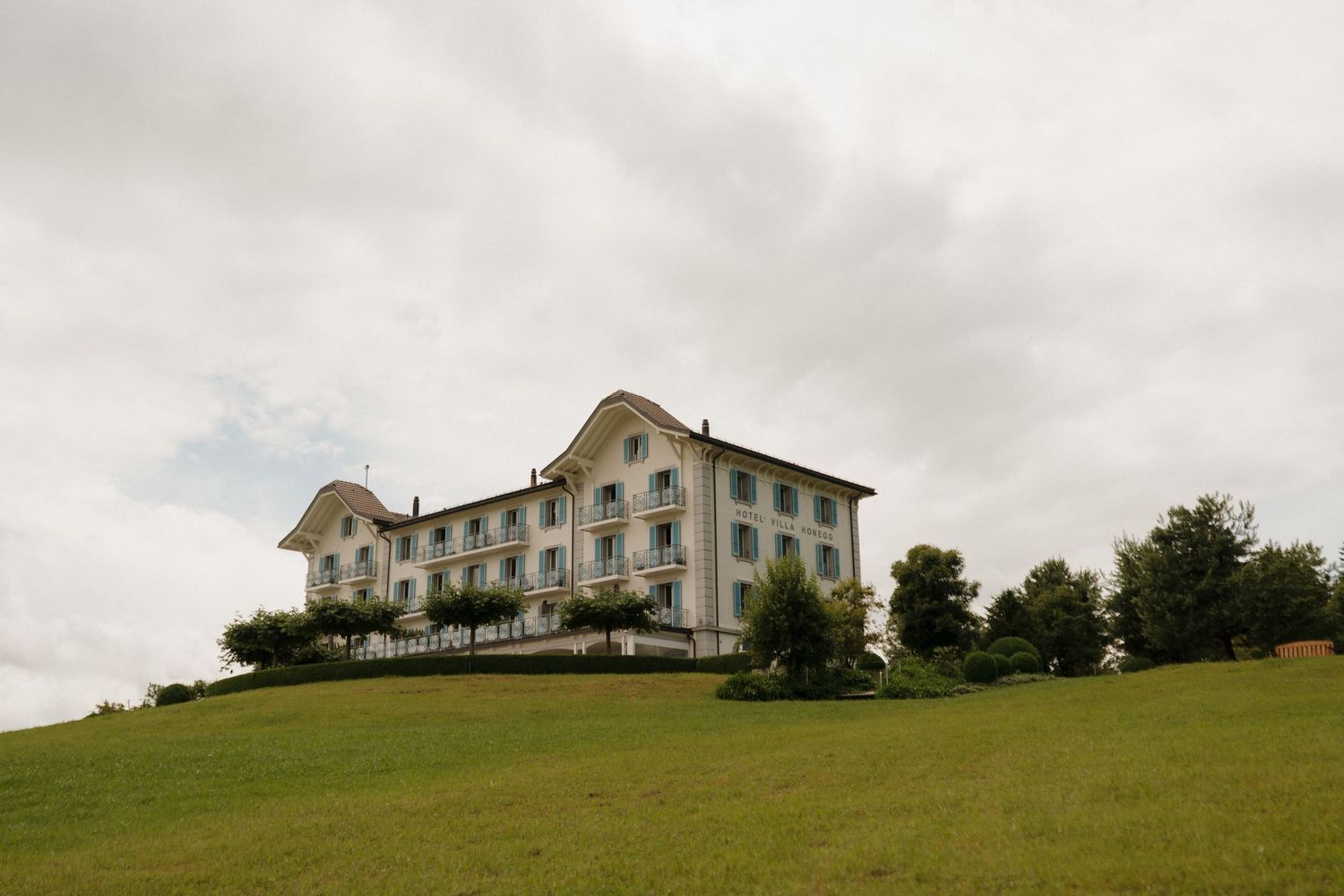 Grand historic Swiss villa sits atop a green hillside under an overcast sky with manicured lawns.