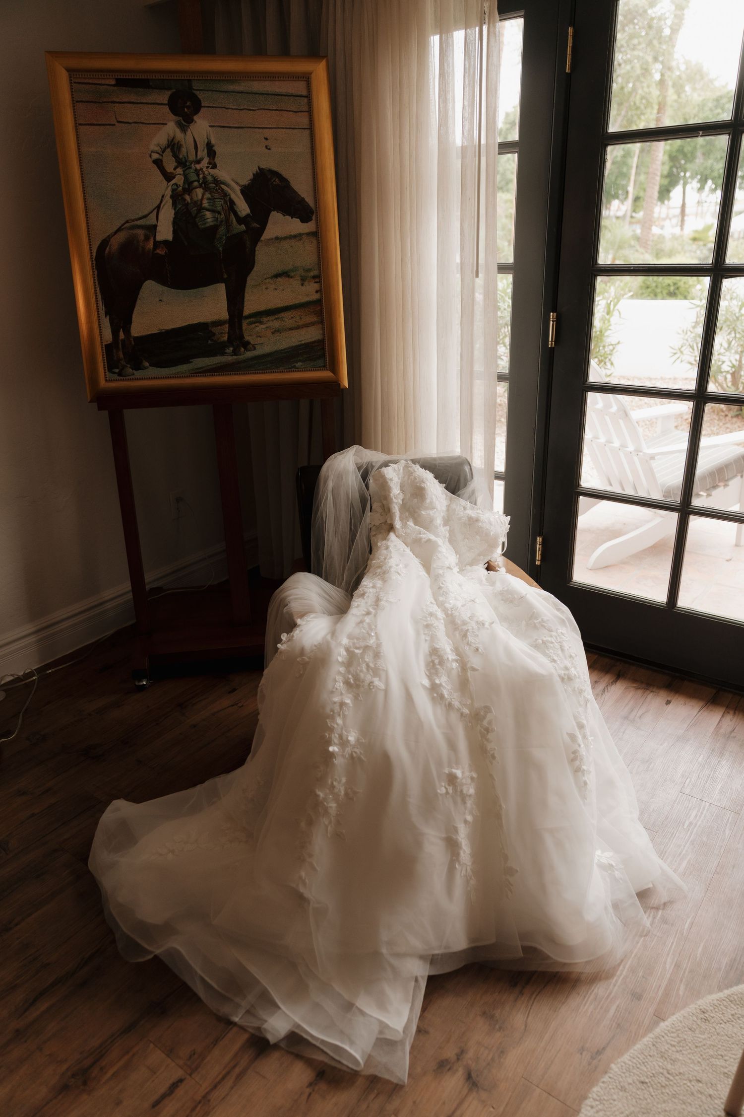 Elegant lace wedding gown draped over chair near French doors with equestrian painting.
